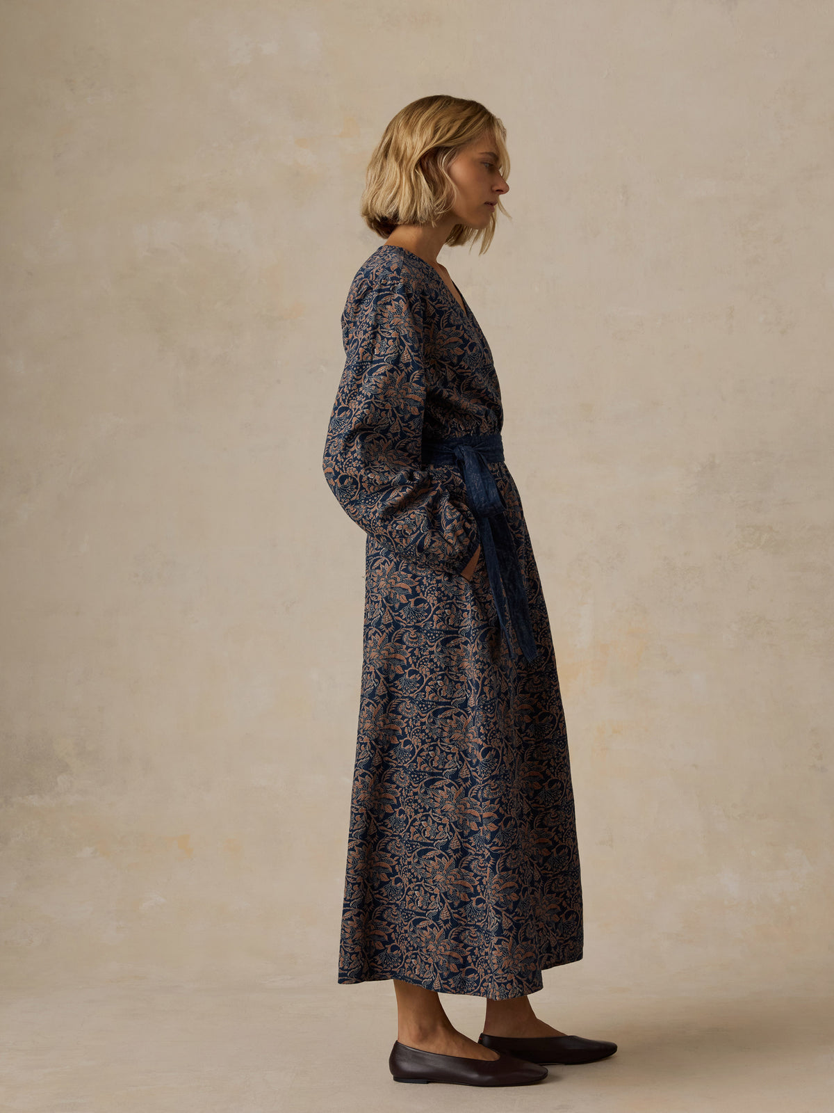 A woman wearing a dark navy and brown floral print shirt dress with three-quarter sleeves, a button-down collar, and a drawstring waist, posed against a textured beige studio backdrop., image 5 of 5
