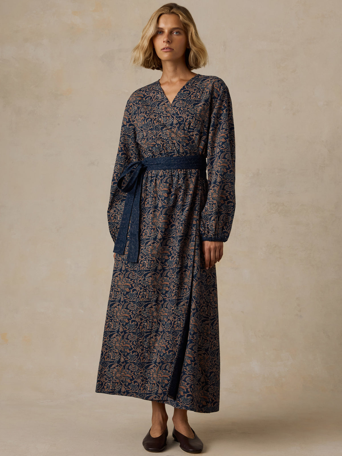 A woman modeling a long-sleeve midi dress with a dark navy and brown floral pattern, standing against a textured beige studio backdrop., image 1 of 5