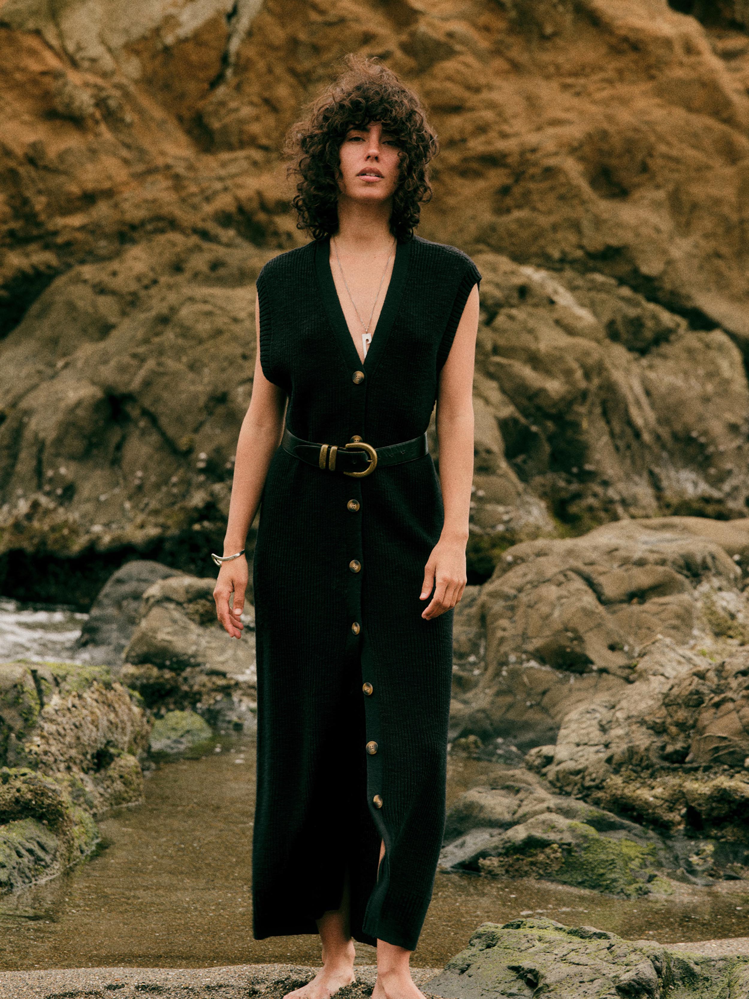 A woman with curly dark hair stands barefoot on rocky coastal ground wearing a sleeveless black maxi dress with a V-neck, front button closure, and a matching belt with a large round buckle.