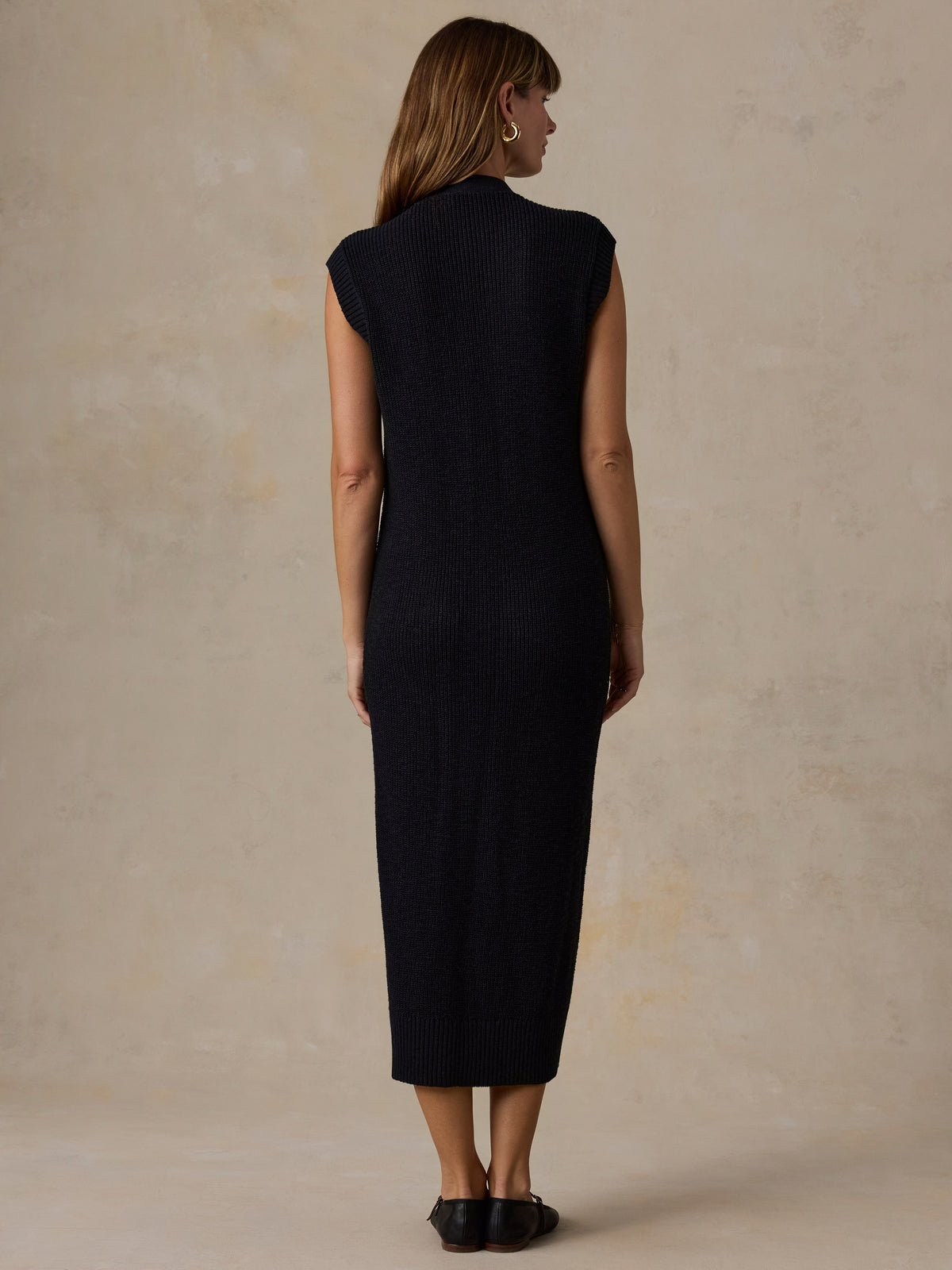 A woman viewed from the back wearing a sleeveless, high-neck, knee-length black ribbed knit dress, standing against a textured beige studio backdrop., image 5 of 5