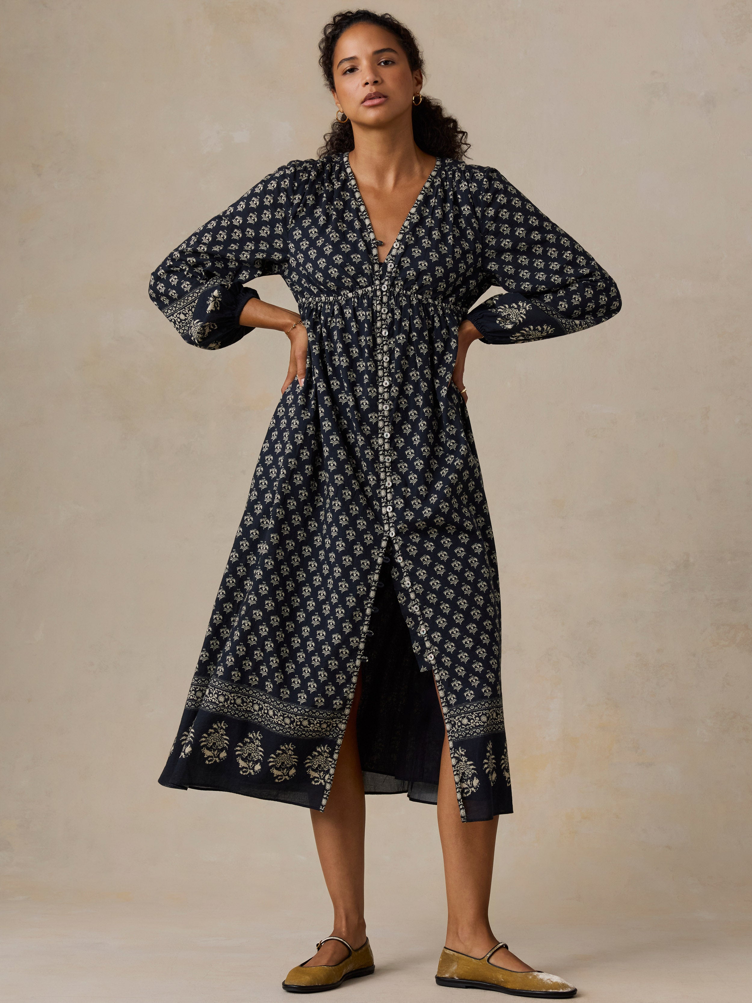 A woman modeling a long navy blue midi dress with a white geometric pattern and a matching belt, standing against a textured beige studio backdrop.