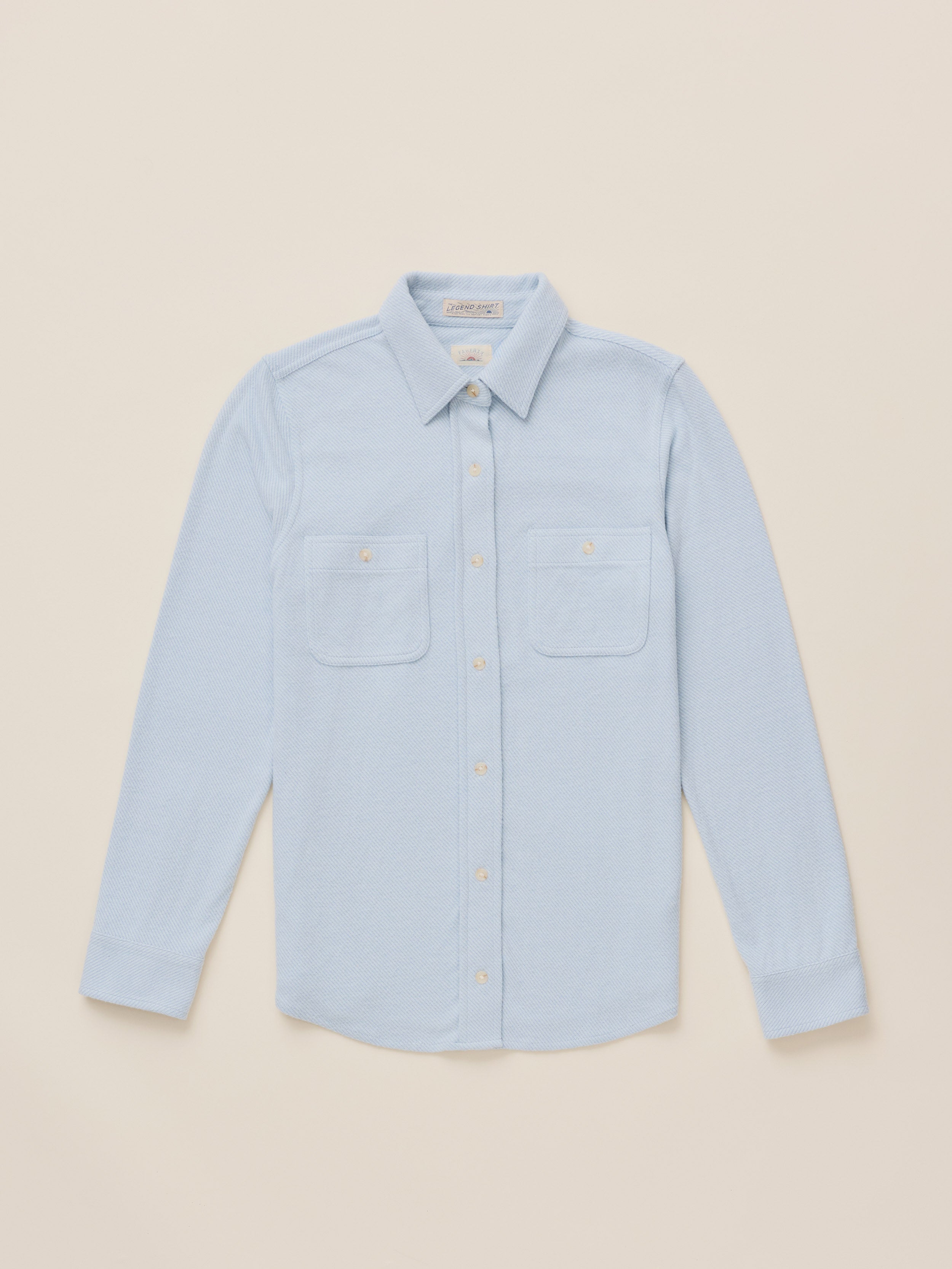 A light blue long-sleeve button-up shirt with two chest pockets, laid flat on a warm beige background.
