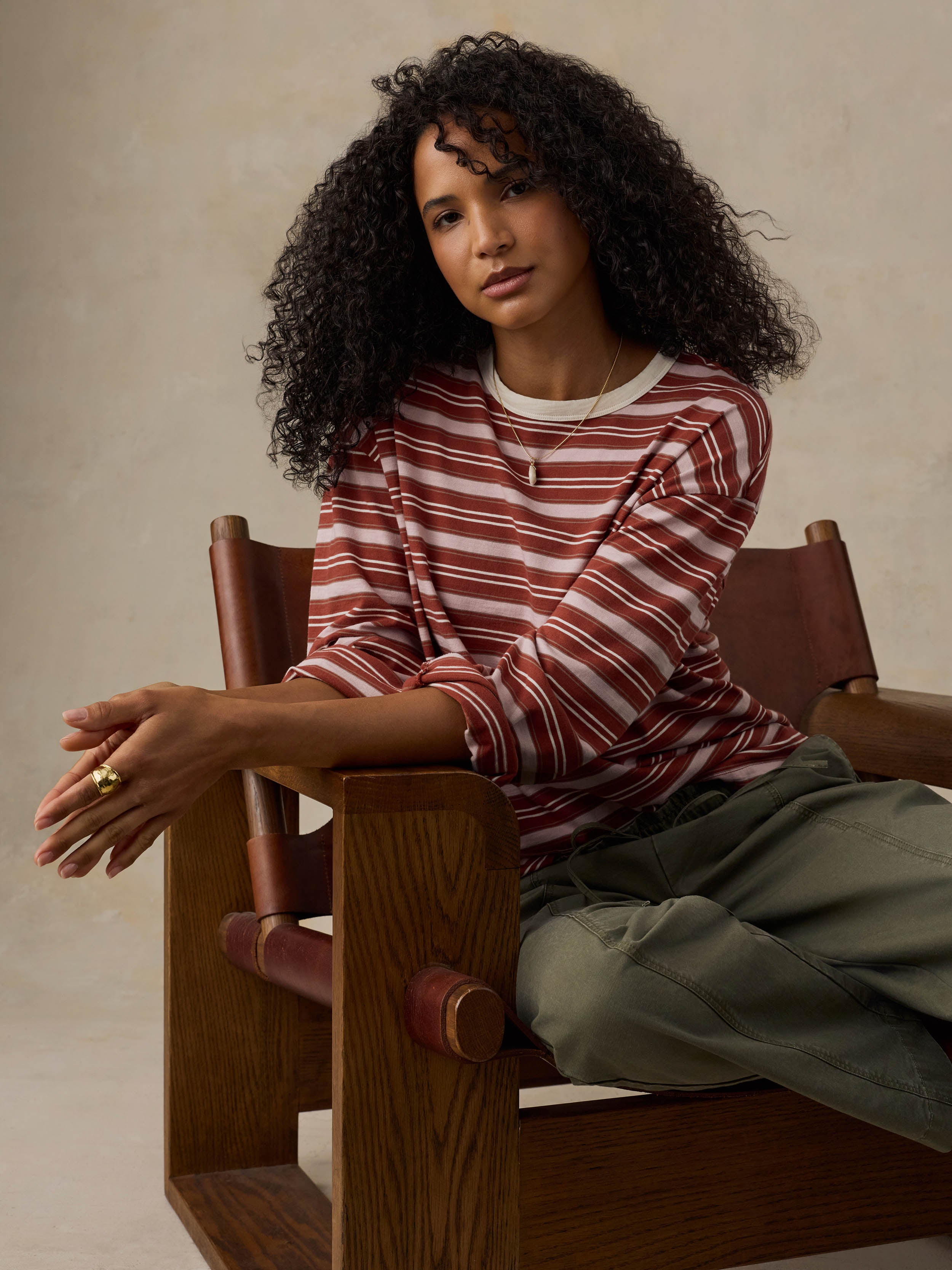 Woman sitting on a wooden chair wearing a red and pink striped long-sleeve shirt and green pants against a beige background