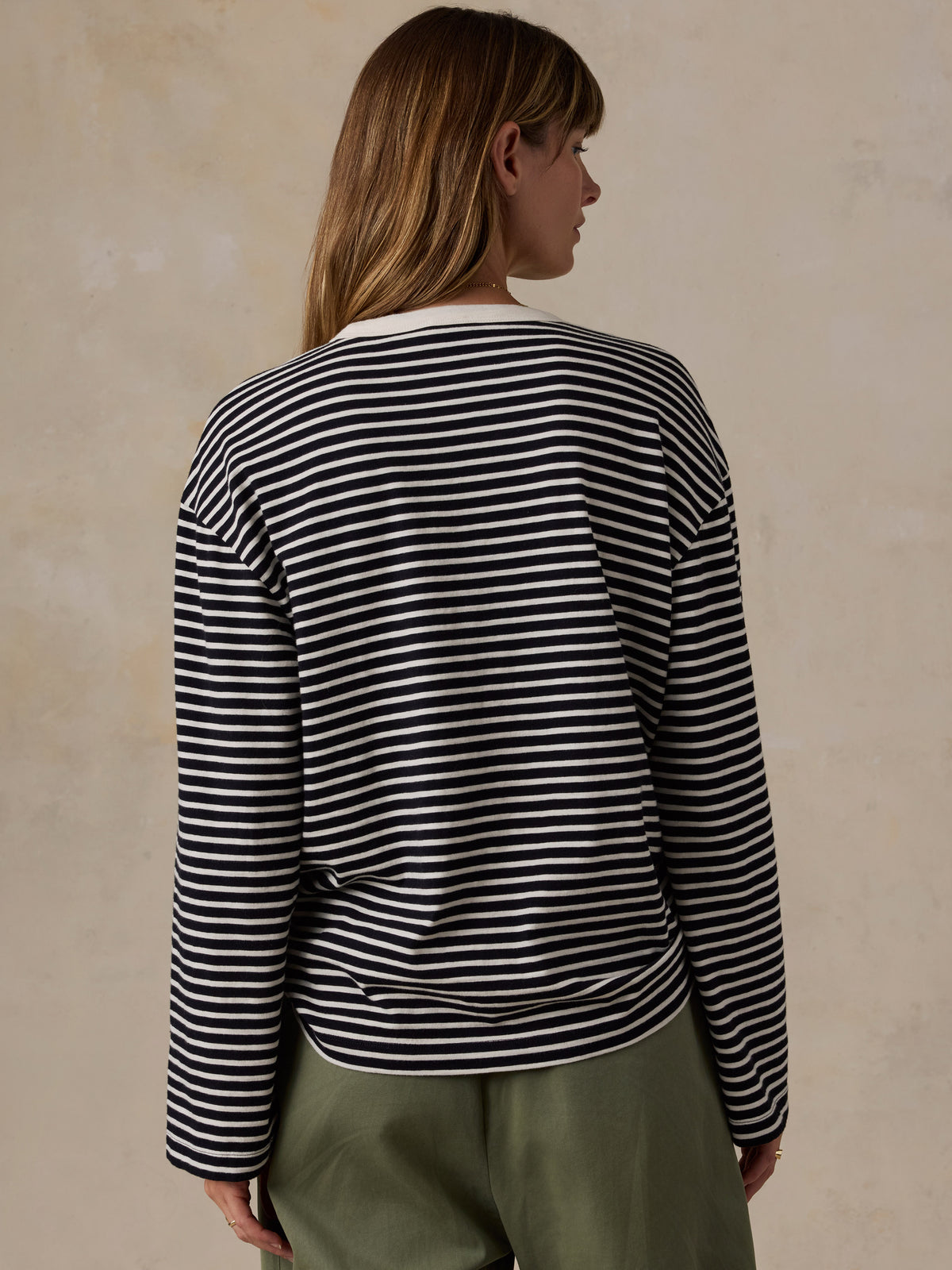 A woman shown from the back wearing a black and white horizontally striped long-sleeve top and olive green pants, standing against a textured beige wall., image 6 of 9
