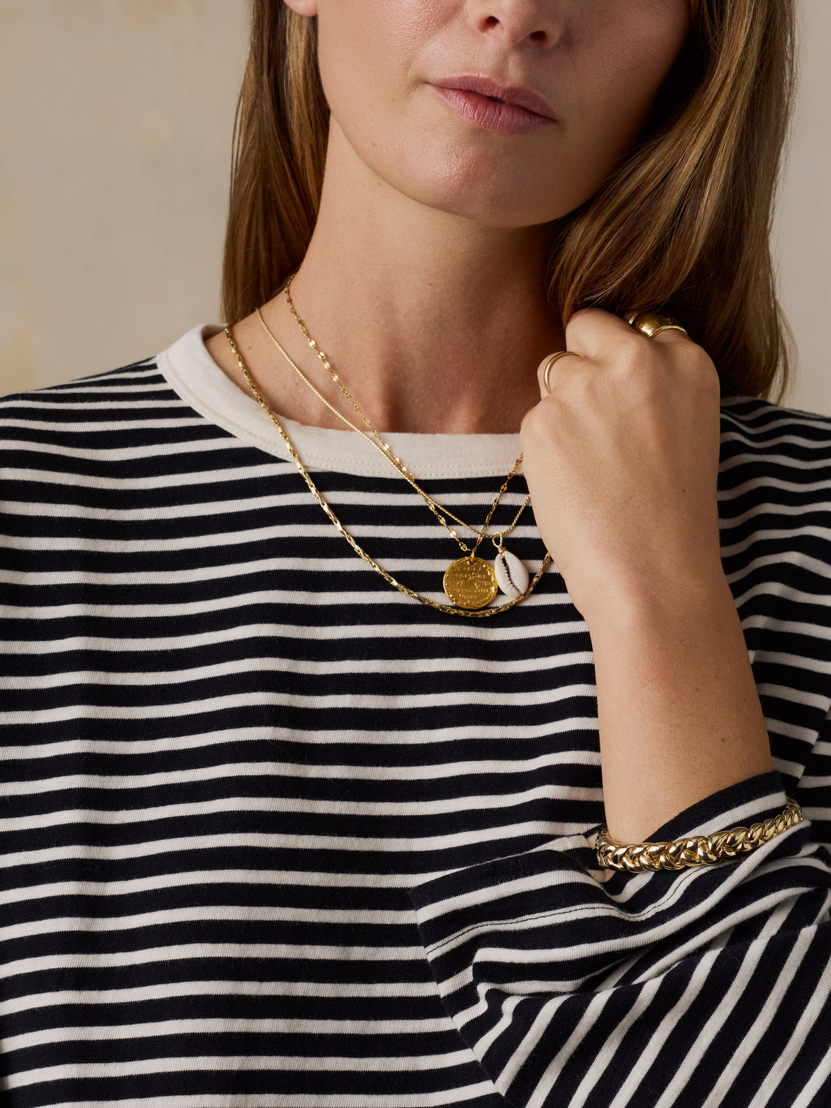 A close-up of a woman wearing a black and white horizontally striped long-sleeve top, layered with gold necklaces, a gold ring, and a gold chain bracelet., image 7 of 9