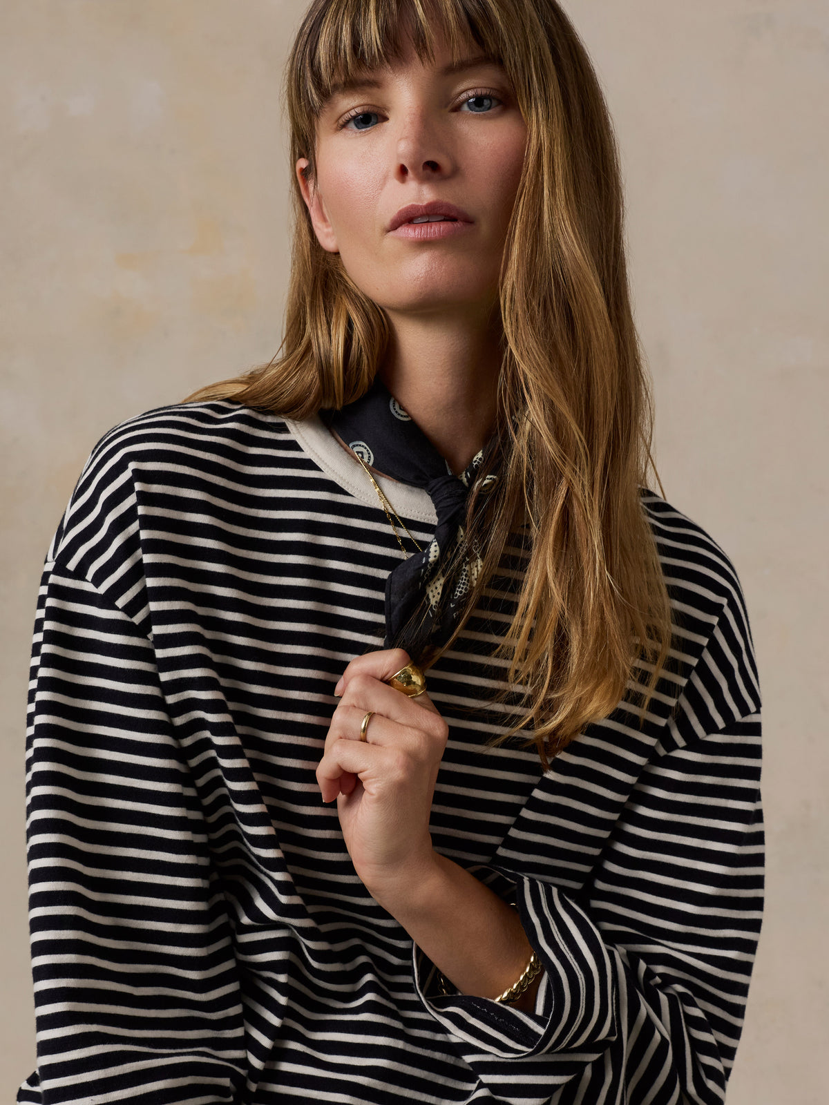 A woman with long light brown hair wearing a black and white horizontally striped long-sleeve top, styled with a dark patterned neck scarf and gold jewelry, set against a textured beige background., image 4 of 9