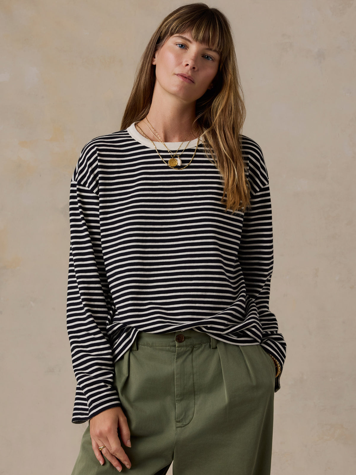 A woman wearing a black and white horizontally striped long-sleeve top with olive green trousers, standing against a textured beige background., image 3 of 9