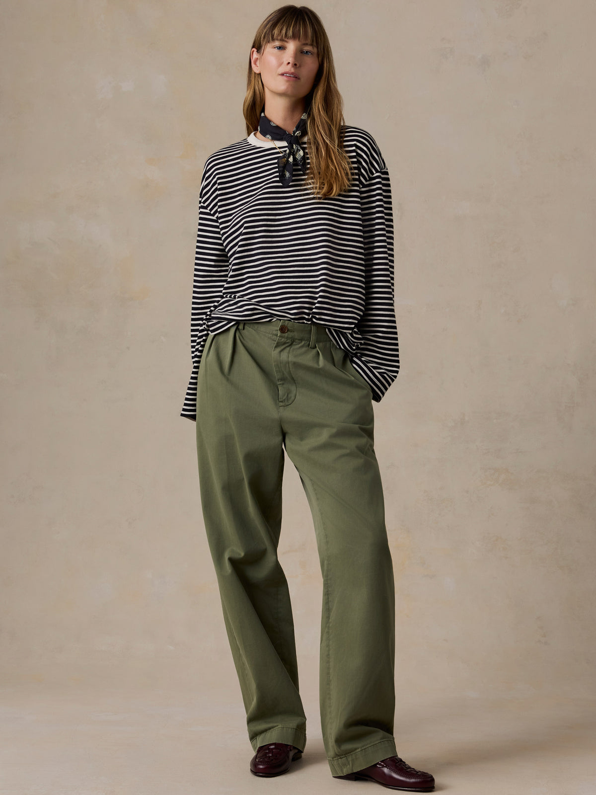 A woman modeling a black and white striped long-sleeve top with a neck scarf detail, paired with olive green wide-leg trousers and dark brown leather shoes, set against a textured beige studio backdrop., image 5 of 9