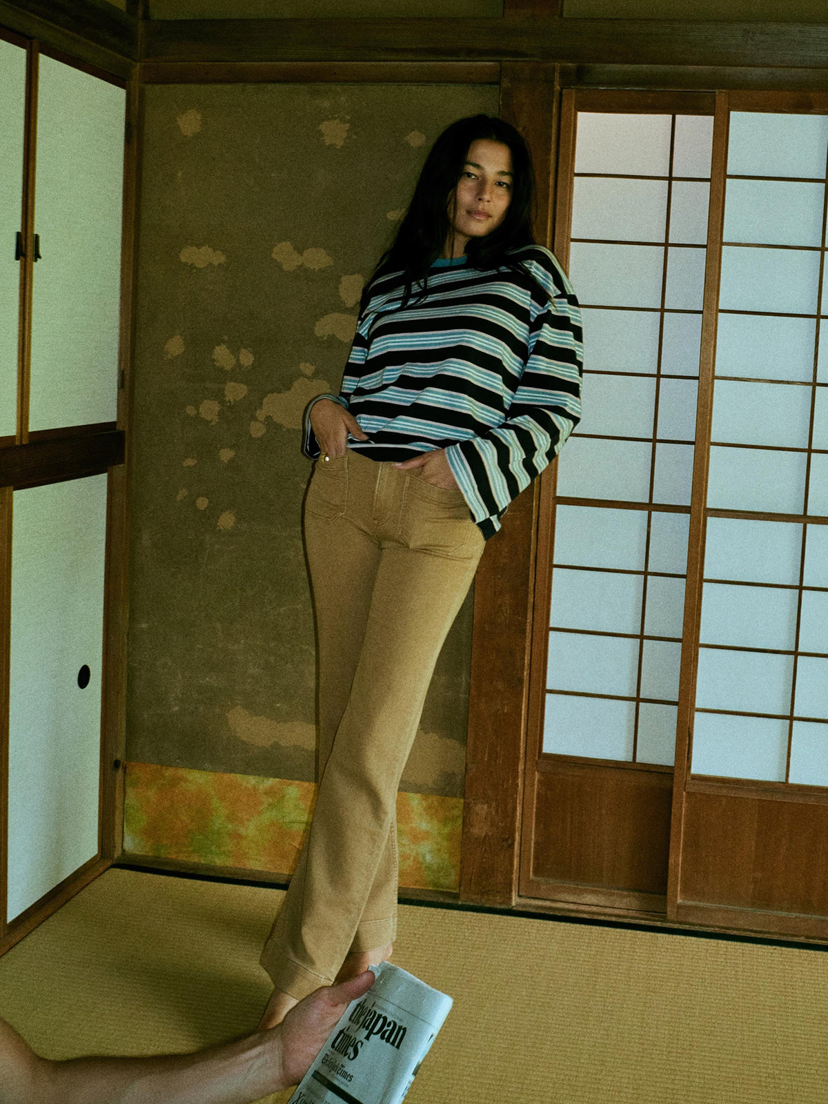 A woman stands in a traditional Japanese room wearing a blue and white striped long-sleeve top and tan pants. A hand in the bottom-left corner holds a newspaper., image 1 of 8