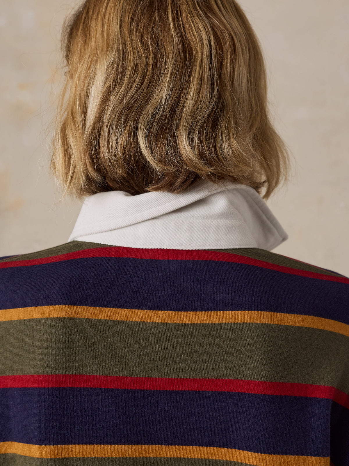 A woman wearing a long-sleeve rugby-style shirt with stripes in olive green, navy blue, red, and mustard yellow, image 6 of 7