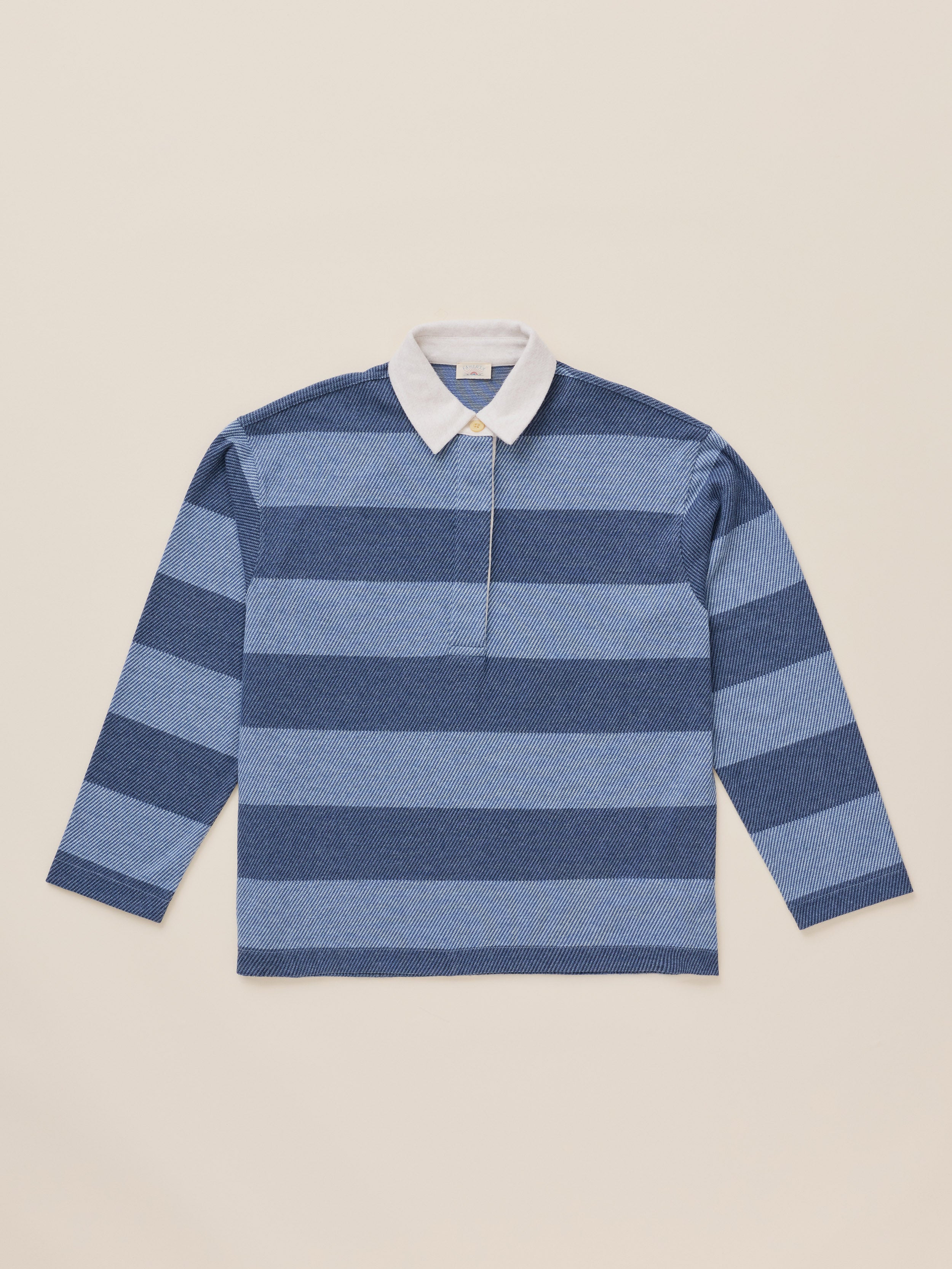 A long-sleeve rugby-style knit polo shirt with horizontal stripes in shades of blue and navy, featuring a white collar, laid flat against a solid beige background.