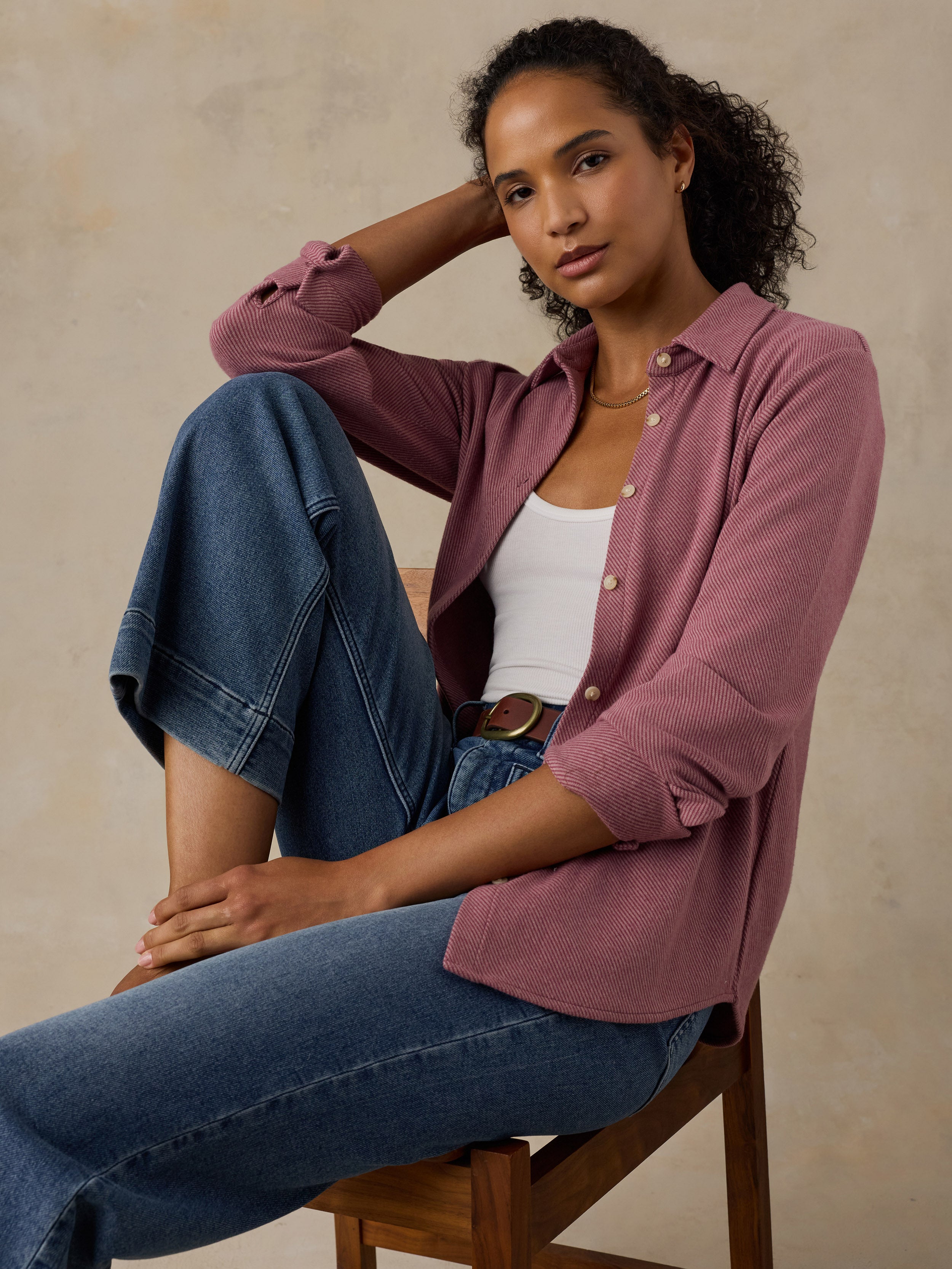Woman sitting on a wooden chair wearing a pink button down shirt and blue jeans against a beige background