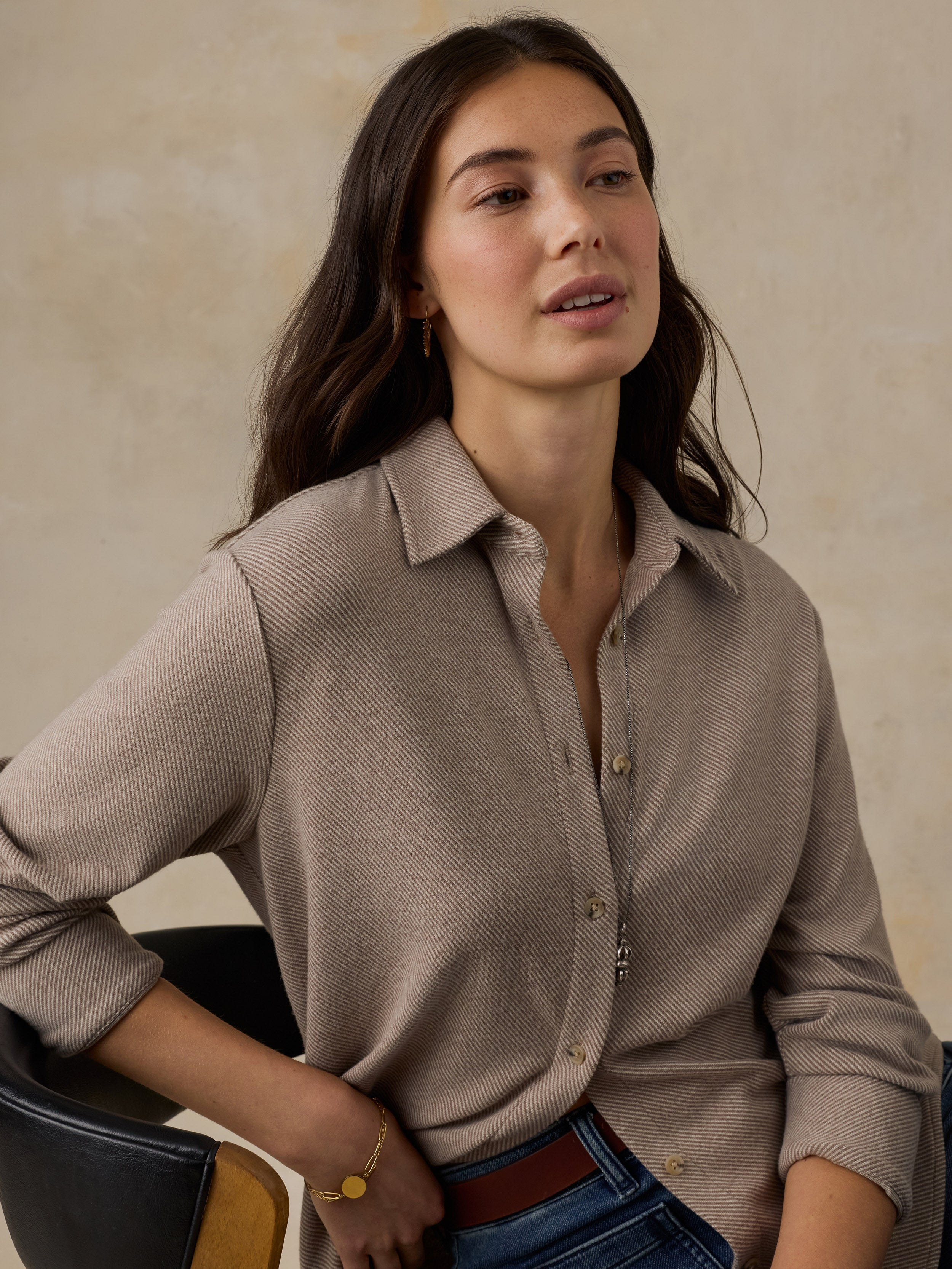 Woman sitting on a wooden chair wearing a beige button down shirt and blue jeans against a beige background