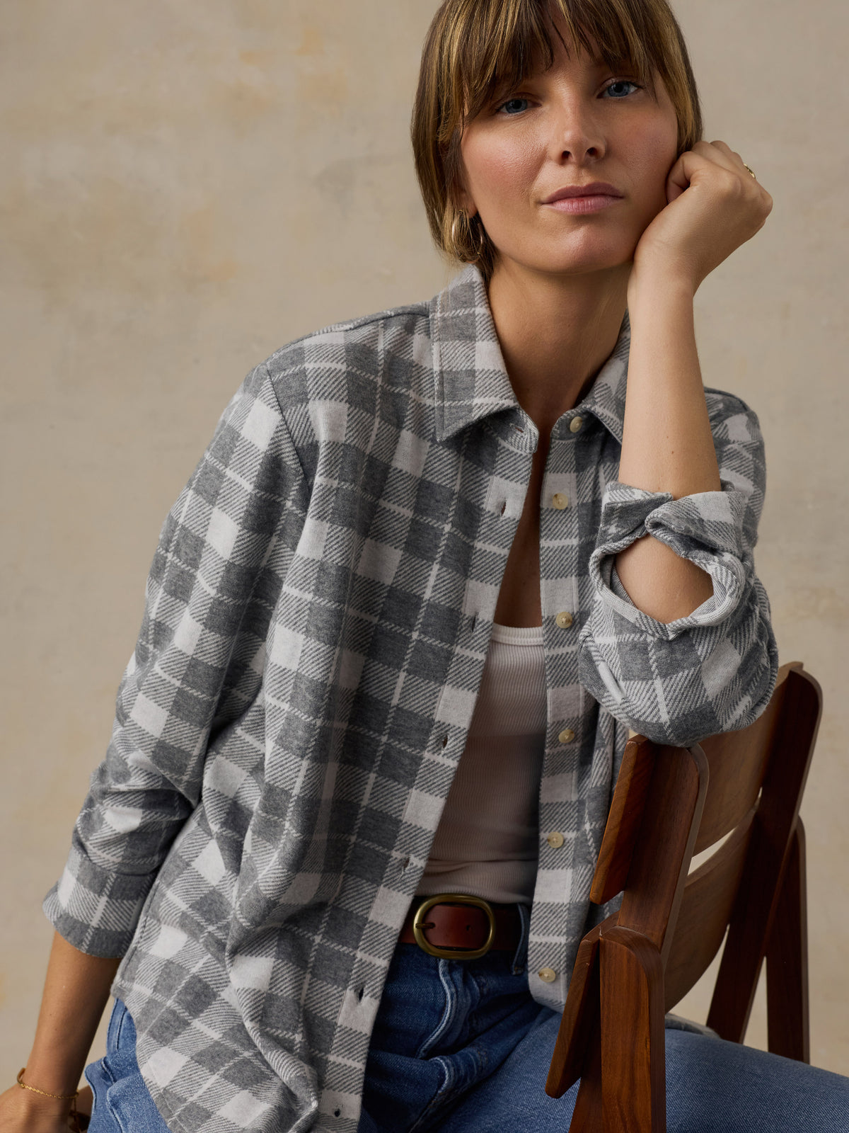 Woman sitting on a wooden chair wearing a grey plaid button down shirt and blue jeans against a beige background, image 2 of 6