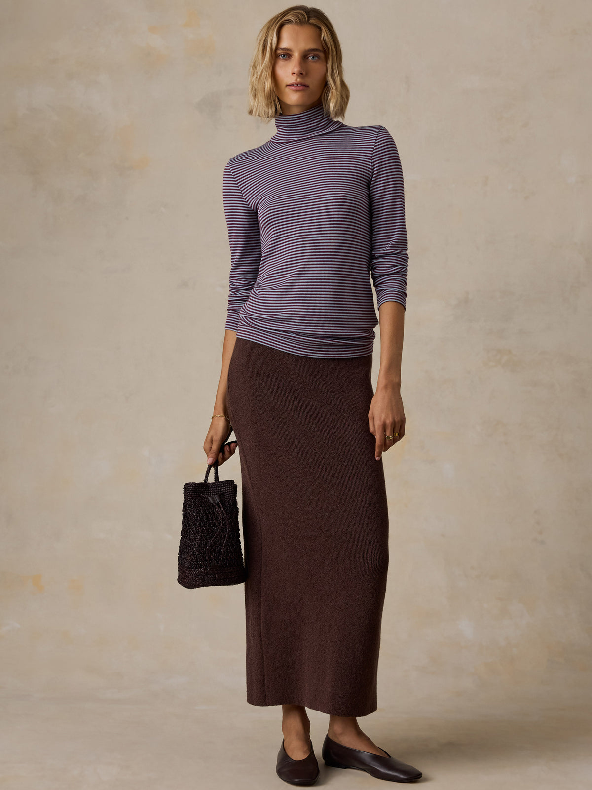 A woman wearing a blue striped turtleneck, brown skirt, and black flats, image 2 of 6
