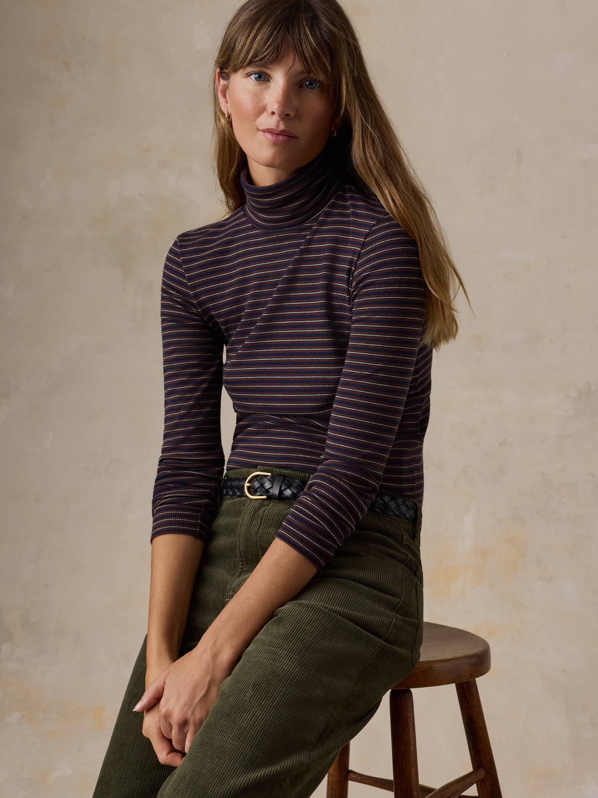 A woman sitting on a stool wearing a brown striped turtleneck and green corduroy pants, image 2 of 6