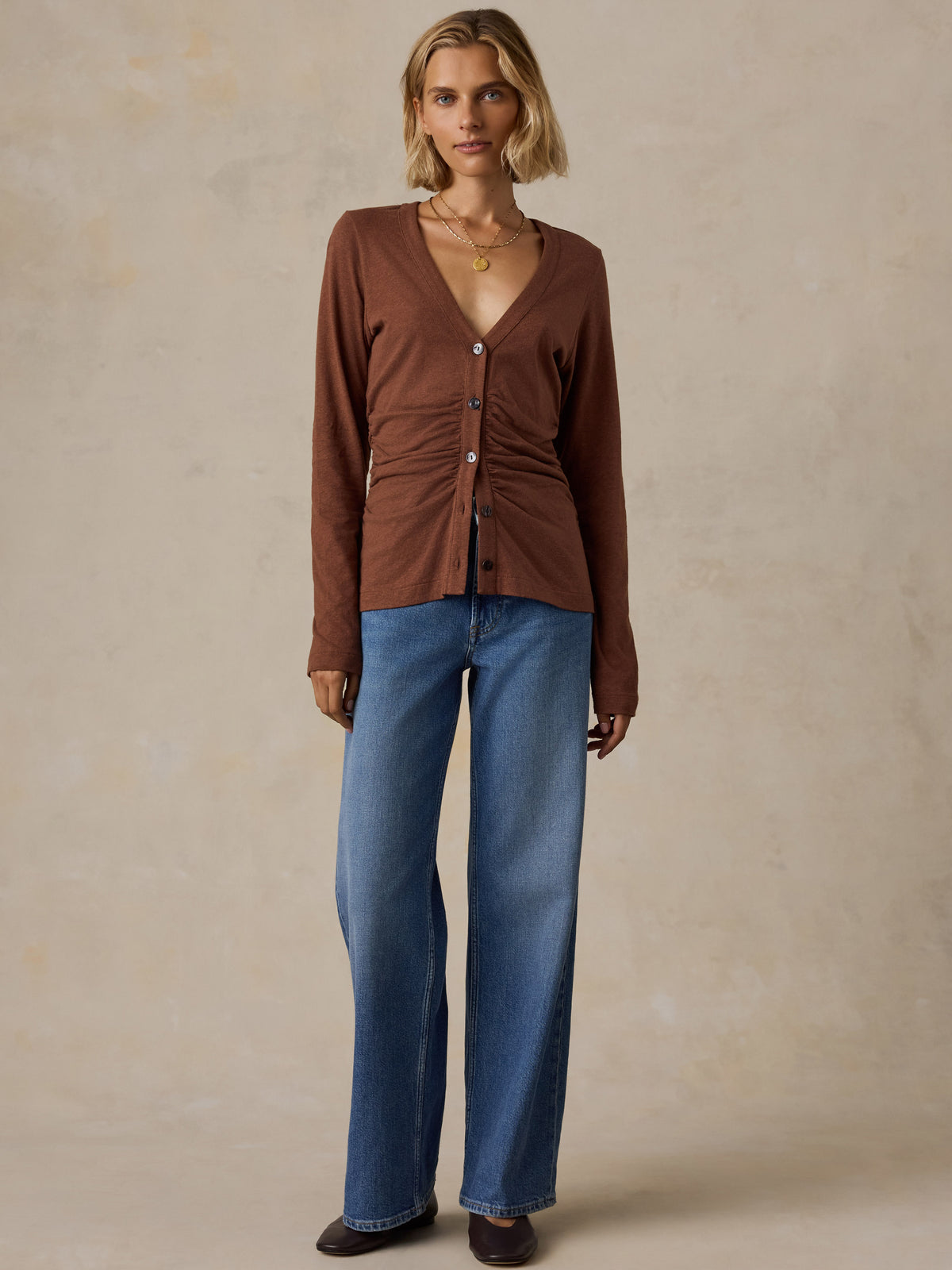 A woman wearing a brown long sleeve top, blue jeans, and black flats, image 2 of 6