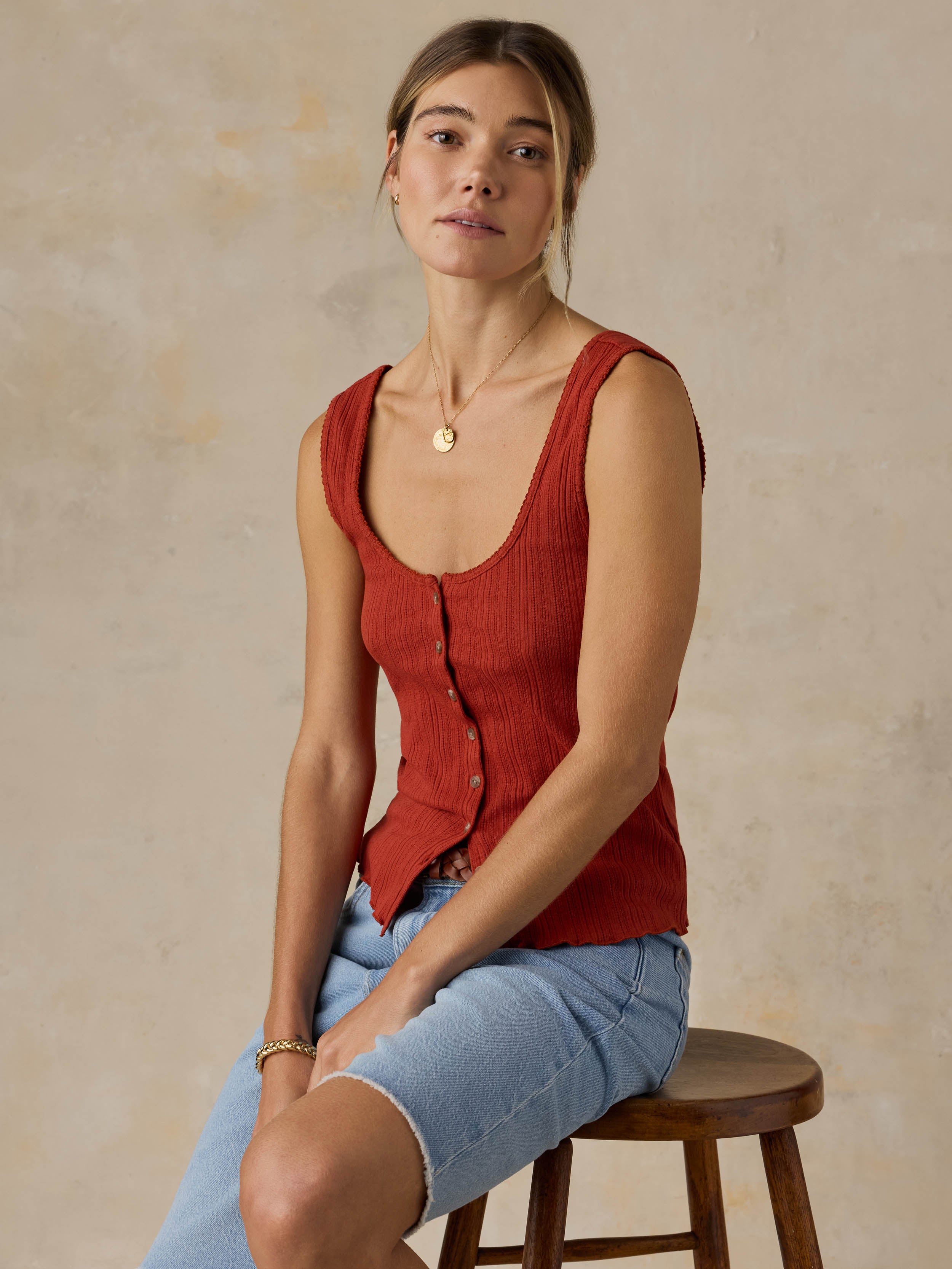 Woman sitting on a wooden stool wearing a red tank top and blue jeans against a beige background