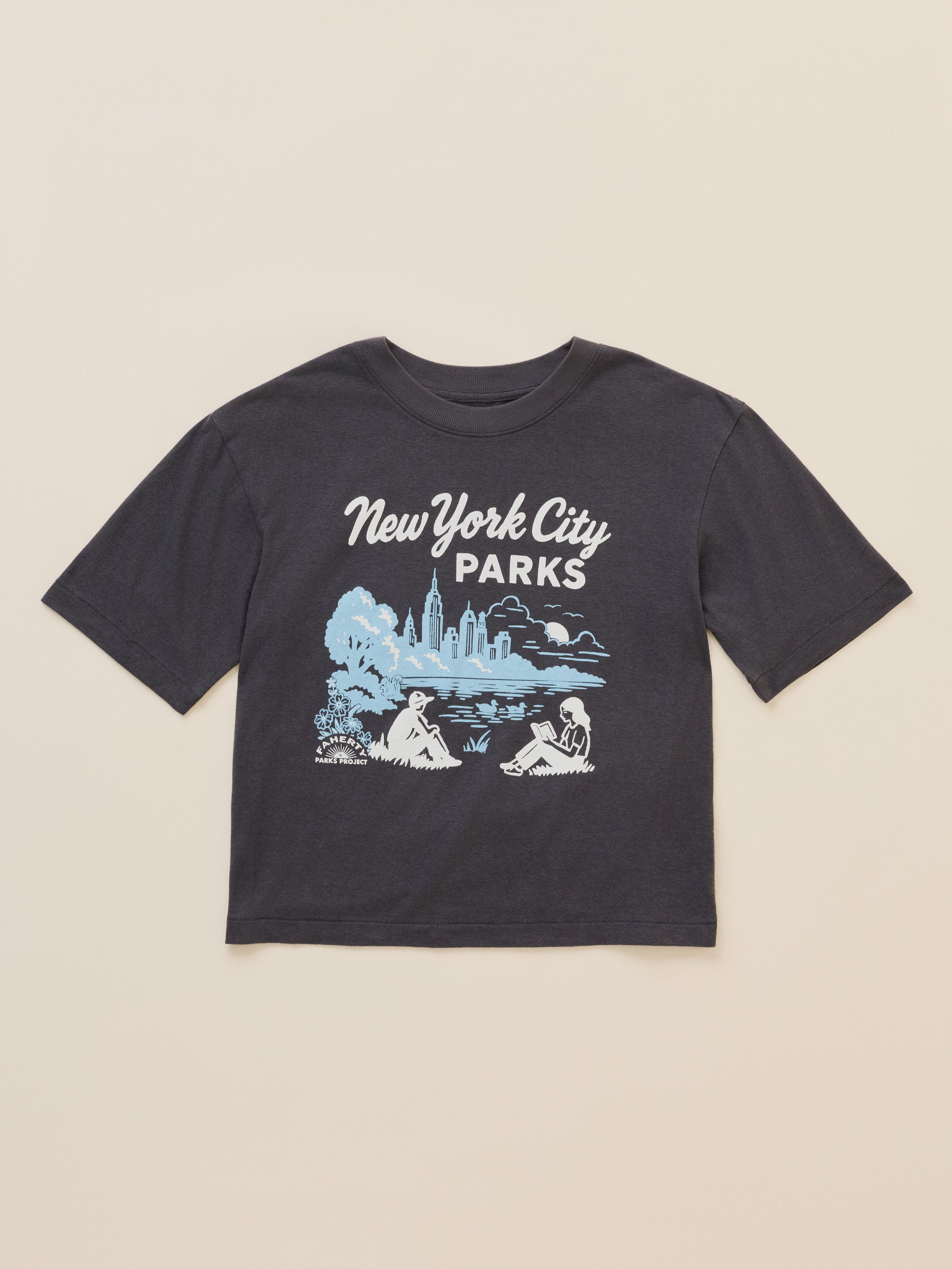 A dark charcoal gray short-sleeve t-shirt with a 'New York City Parks' graphic on the front, featuring a light blue and white illustration of a park scene with a skyline, laid flat against a warm beige background.