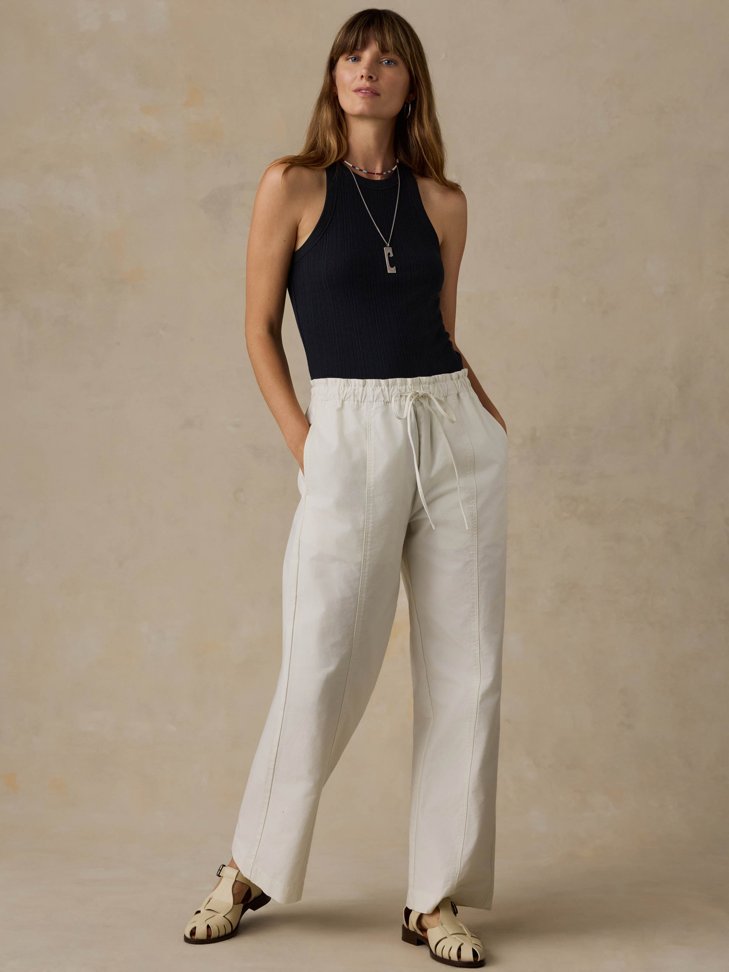 Woman wearing a black tank top and white pants against a beige background
