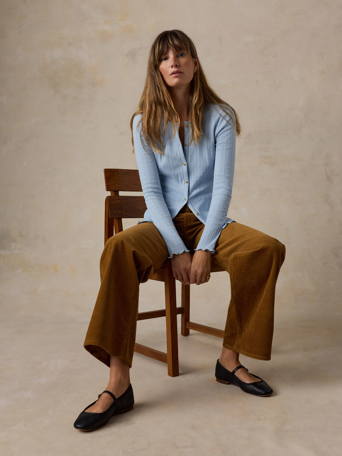 A woman sitting on a chair wearing a light blue cardigan and brown corduroy pants, image 2 of 6
