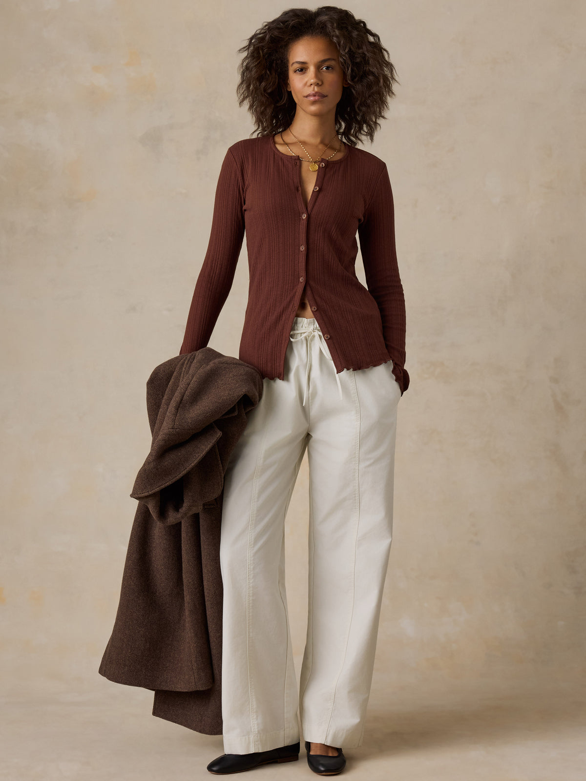 A woman wearing a brown knit cardigan, white pants, and black flats, image 2 of 6