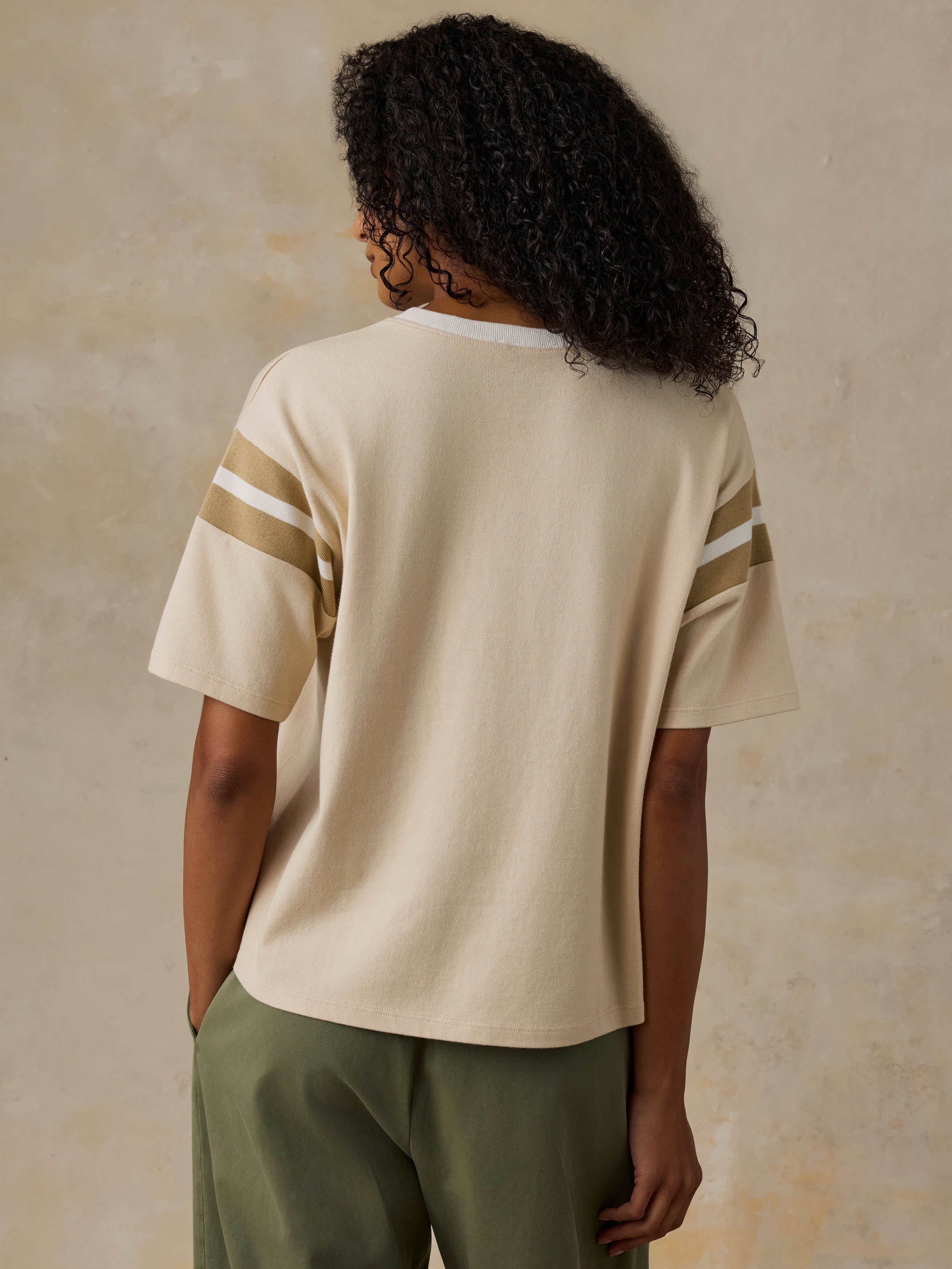 A person woman from the back wearing a cream short-sleeve knit top with two diagonal stripes on each sleeve in a warm tan and white, paired with olive green pants, against a textured beige wall.