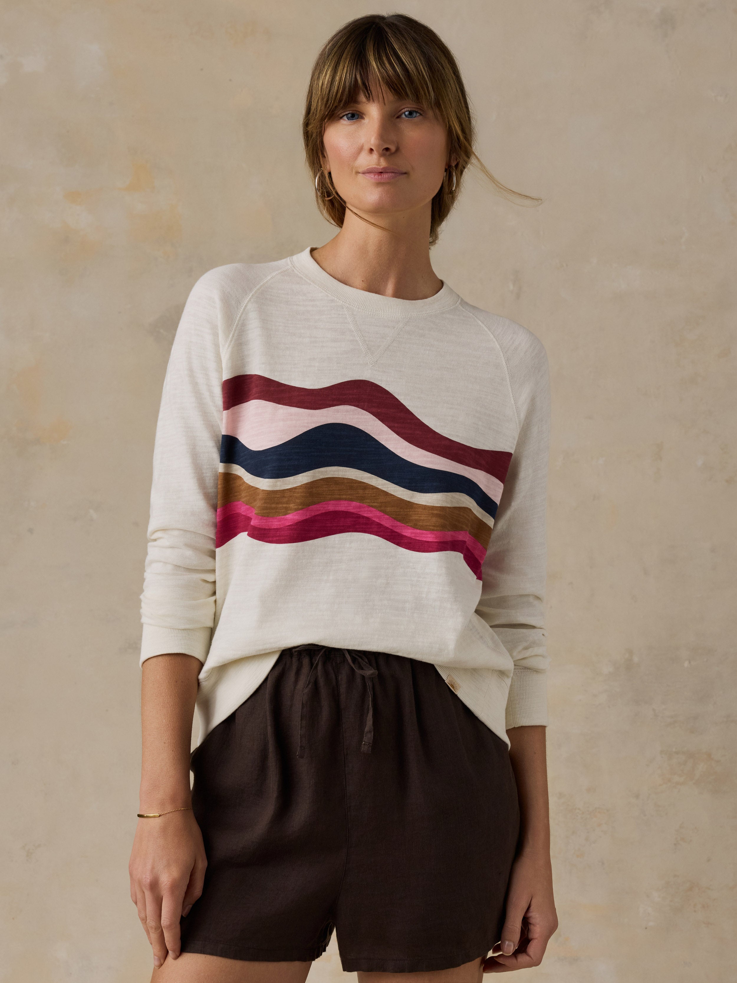 Woman wearing a white and multicolor graphic crewneck with brown linen shorts against a beige background