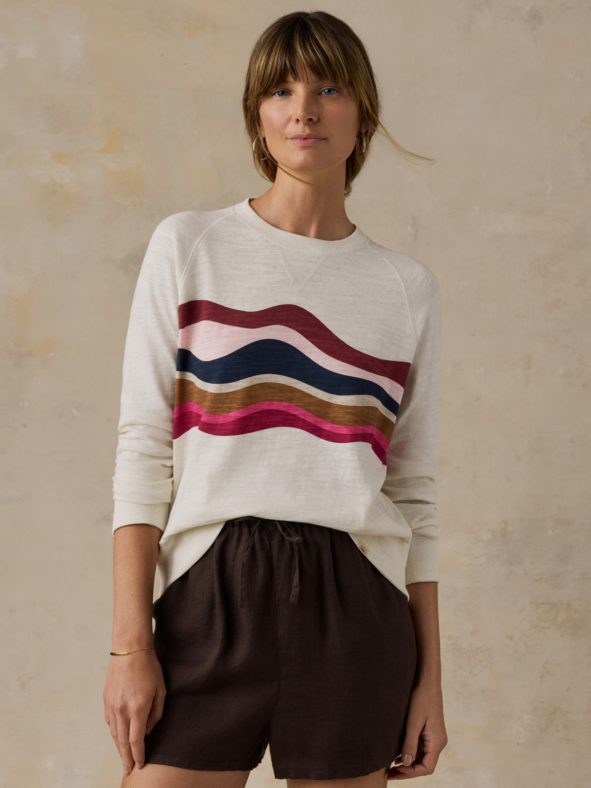 Woman wearing a white and multicolor graphic crewneck with brown linen shorts against a beige background, image 1 of 6