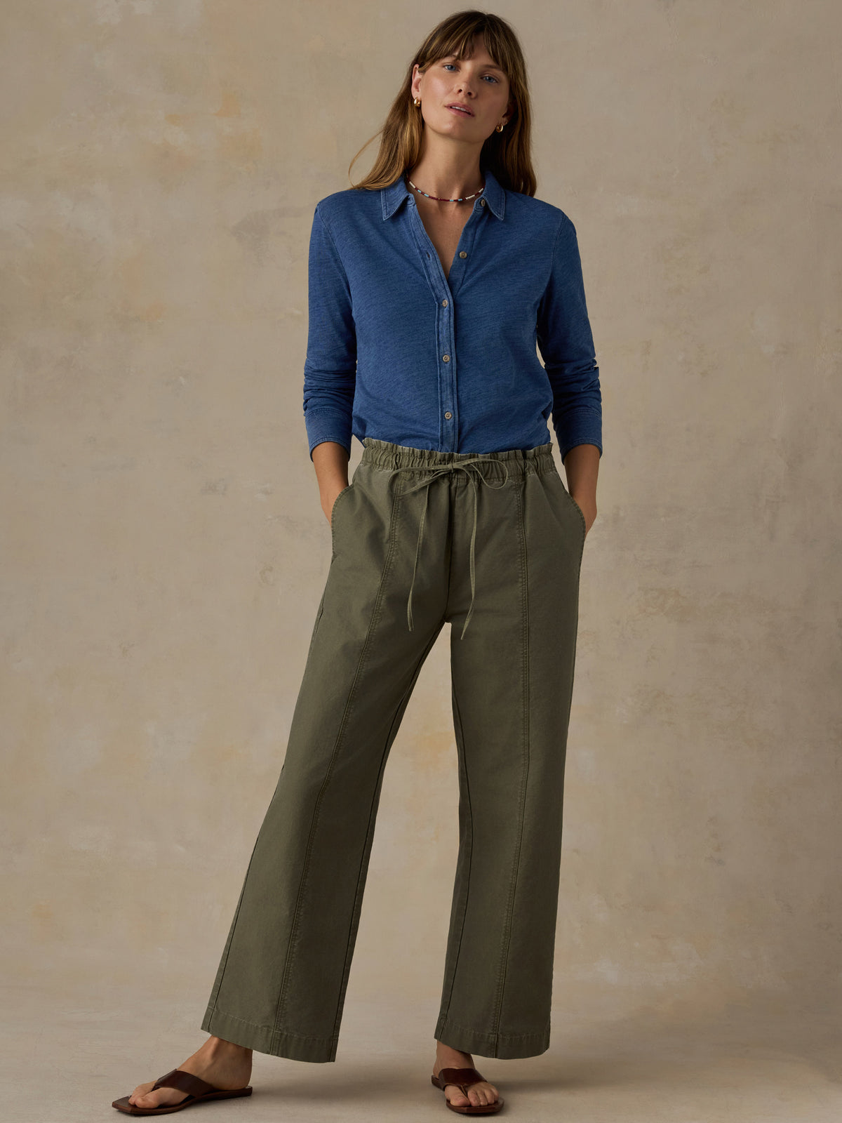 Woman wearing a blue button down shirt and olive green pants against a beige background, image 4 of 6