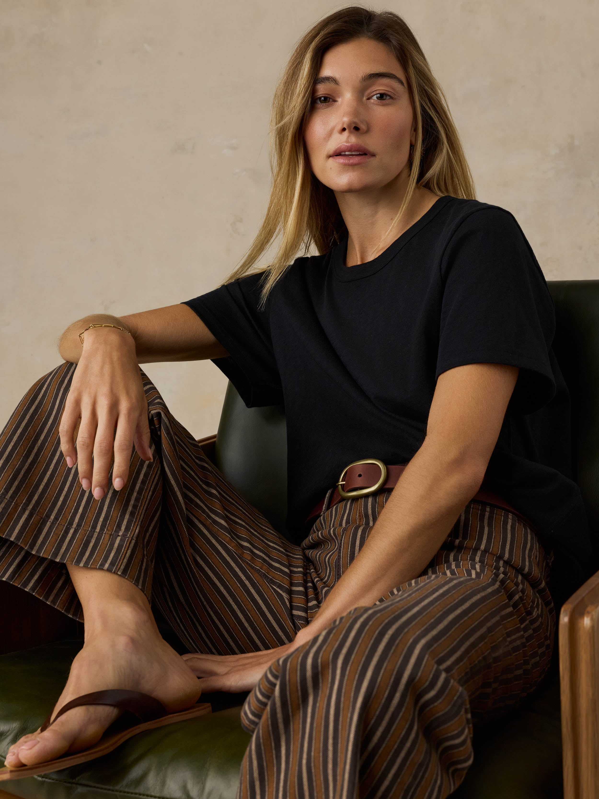 Woman sitting on a wooden chair wearing a black t-shirt and striped pants against a beige background