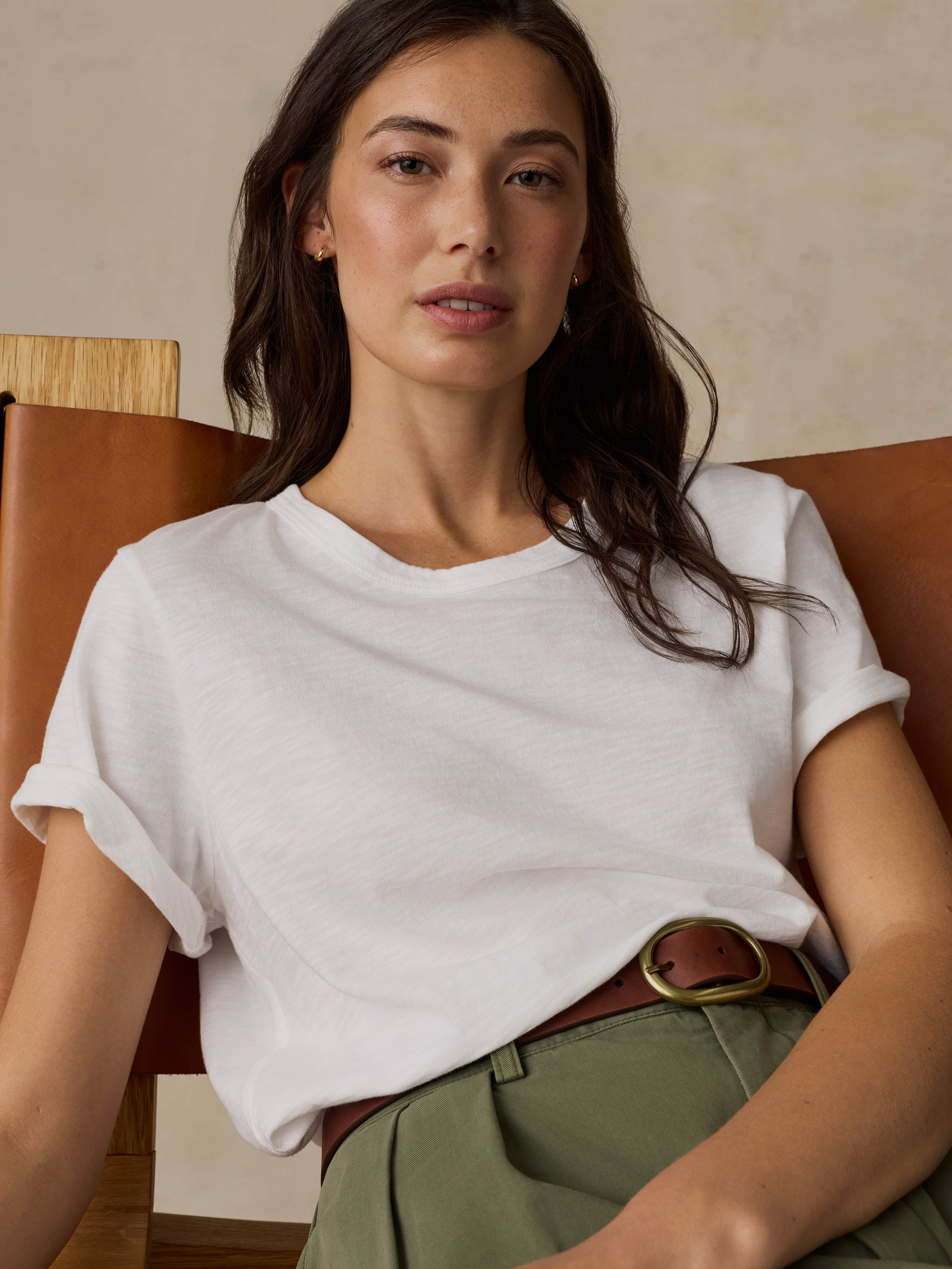 Woman sitting on a wooden chair wearing a white t-shirt and green pants against a beige background