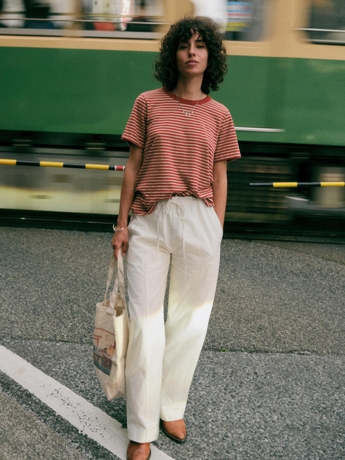 A woman stands on a street in front of a blurred green train, wearing a red and white striped short-sleeve t-shirt, cream wide-leg trousers, and brown shoes, while holding a canvas tote bag., image 1 of 8