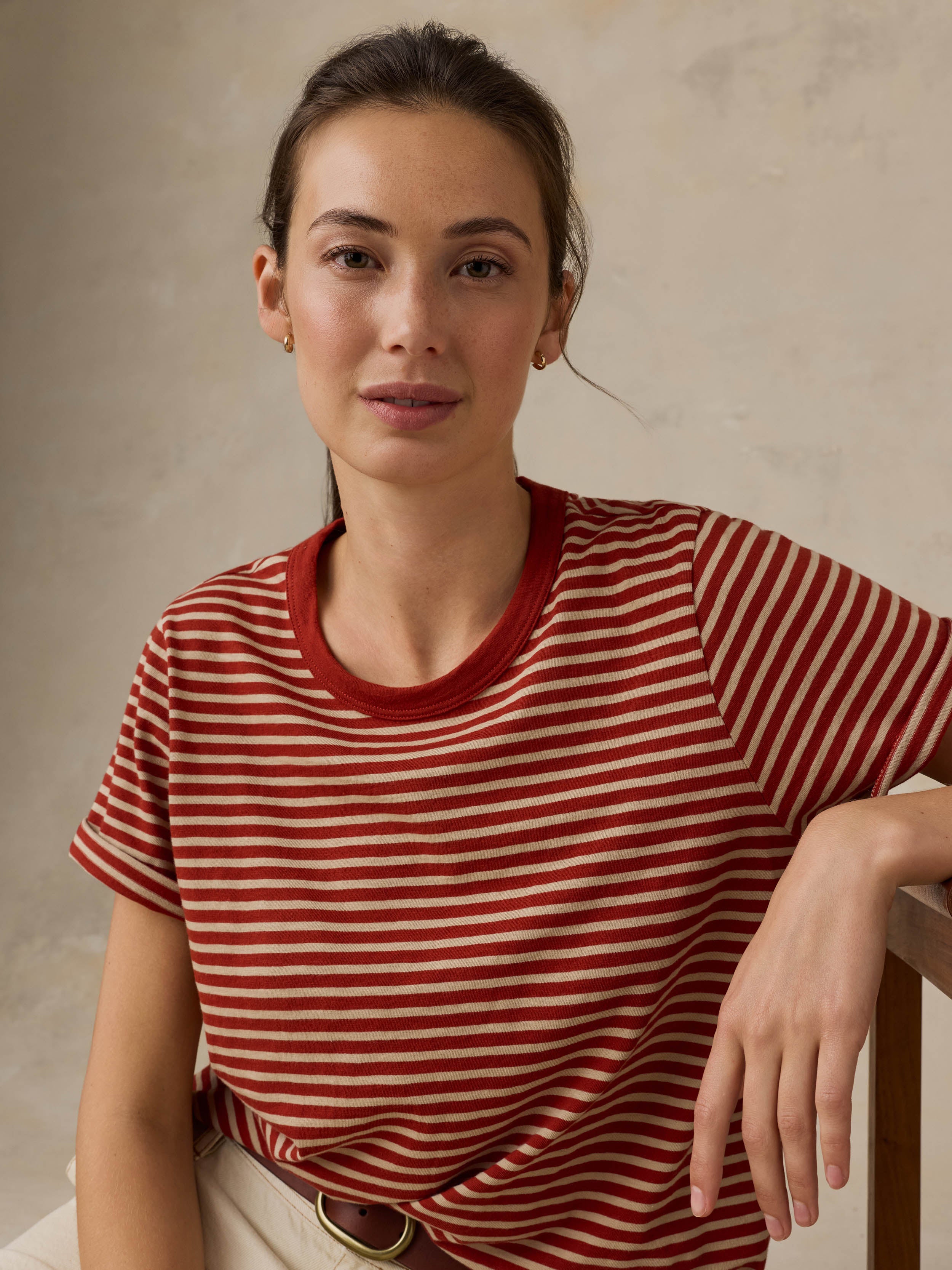 Woman sitting on a wooden chair wearing a red striped shirt against a beige background