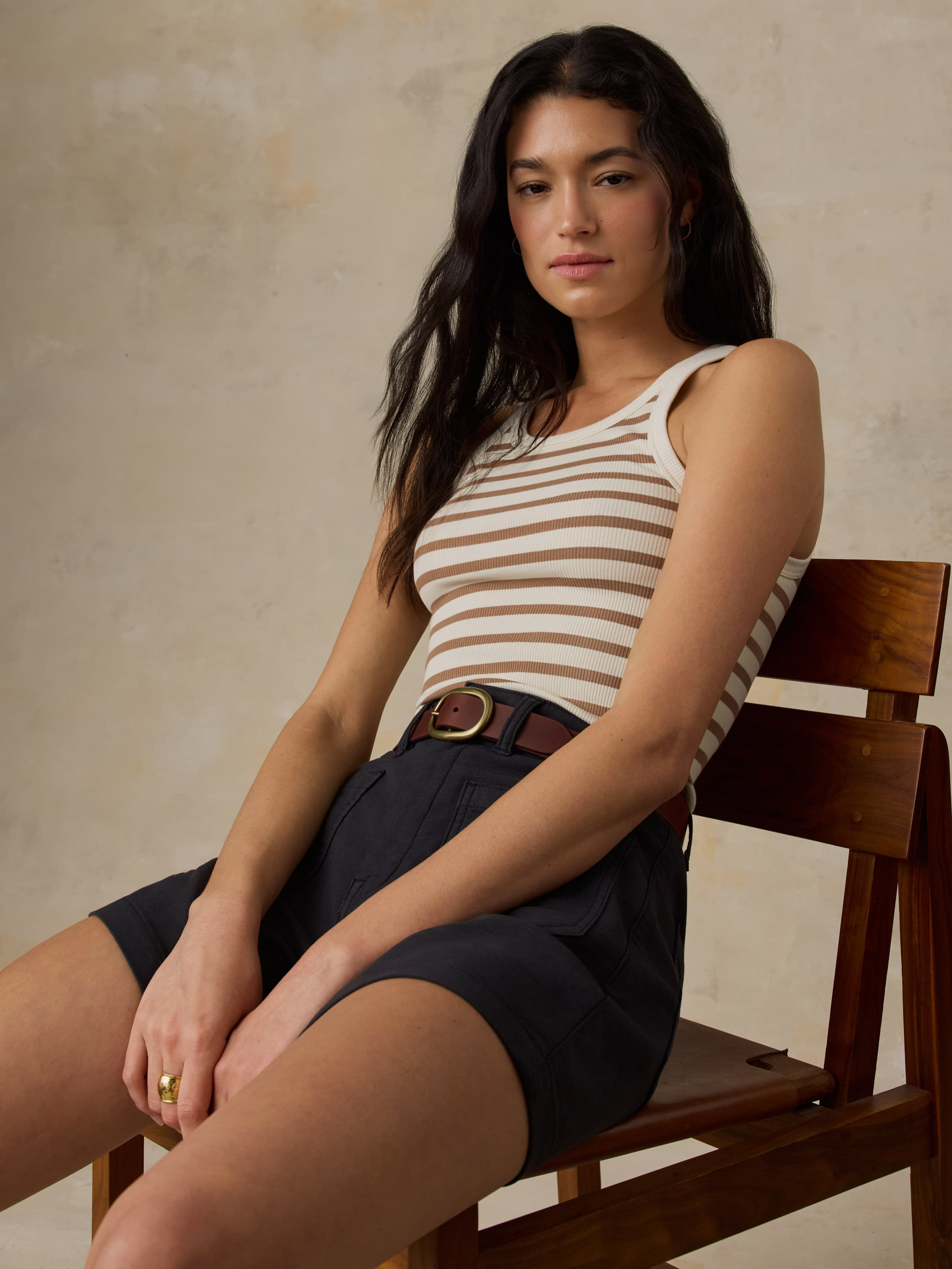 Woman sitting on a wooden chair wearing a brown striped tank top and shorts.