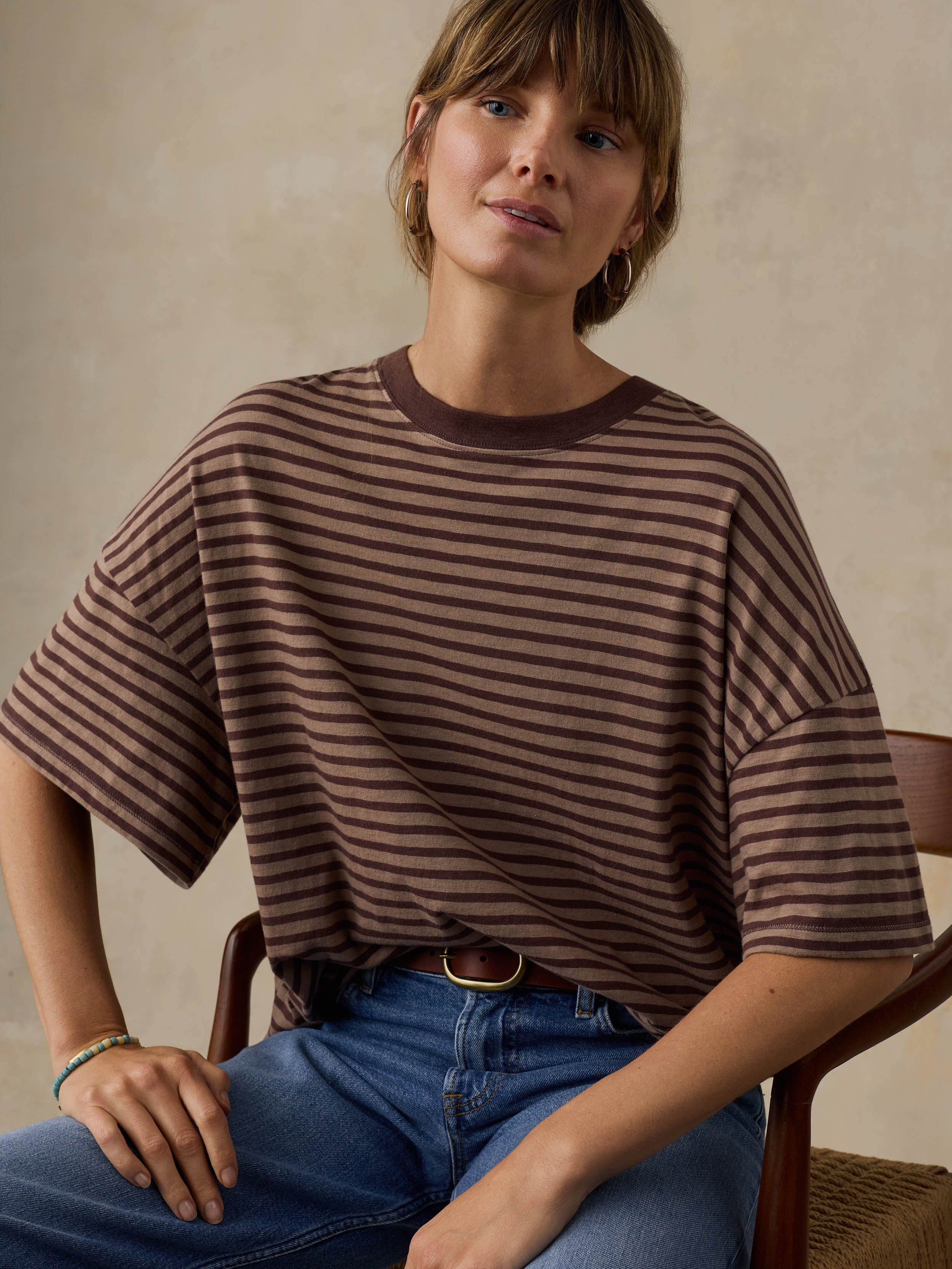 Woman wearing a brown striped sweater and blue jeans, sitting on a wooden chair.