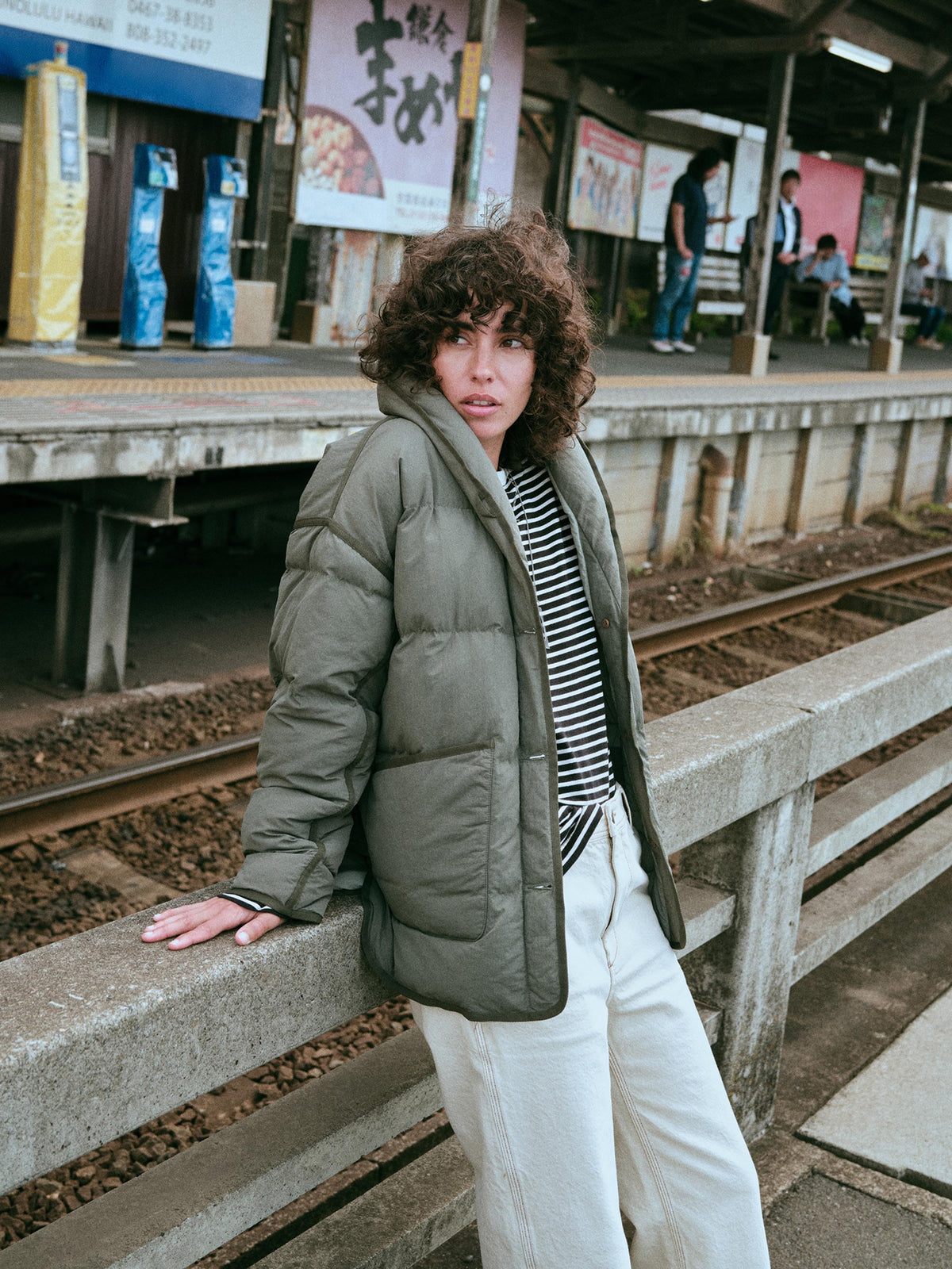 A woman wearing an olive green puffer jacket over a black and white striped shirt and white pants., image 7 of 7