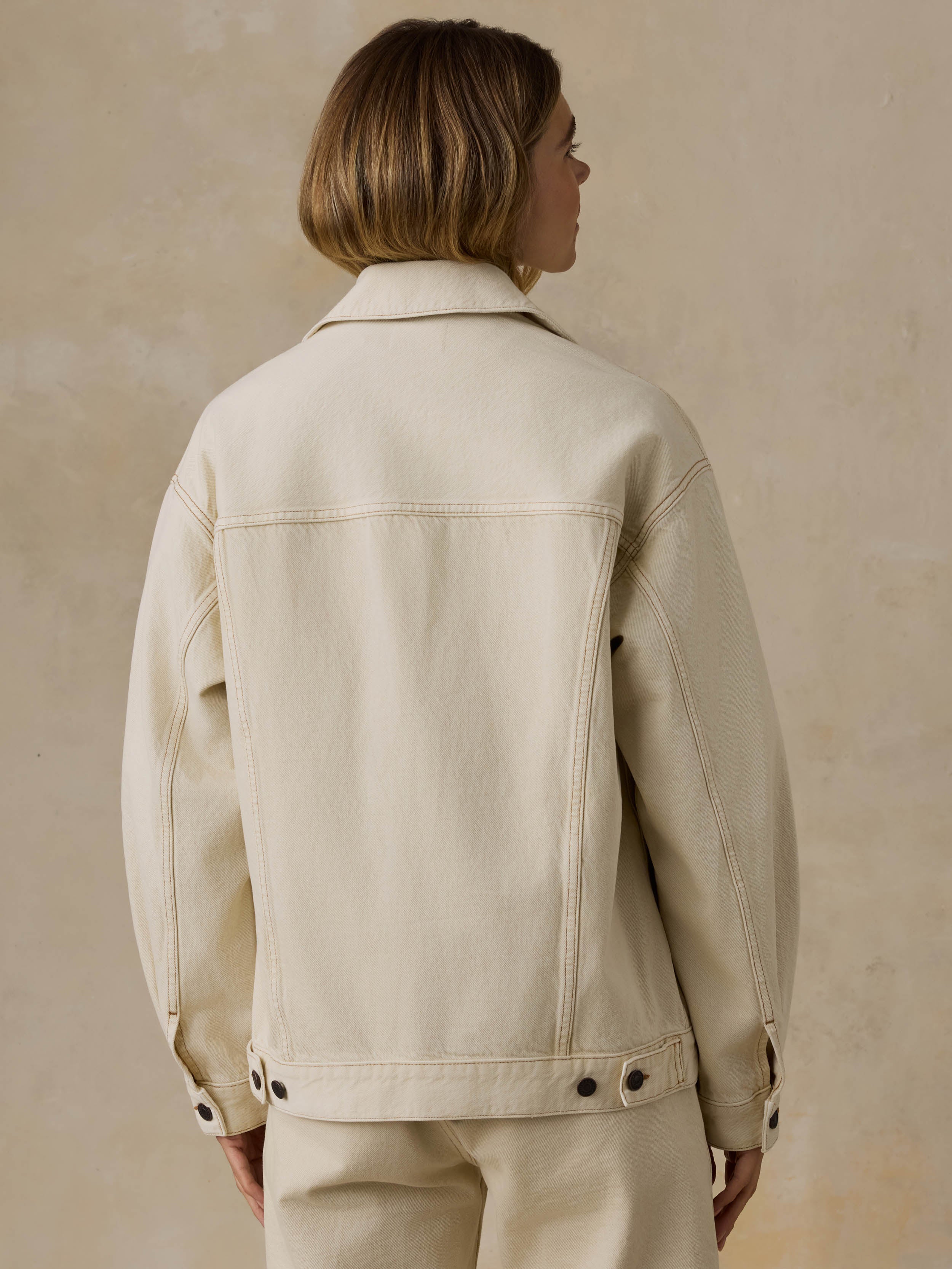Woman wearing an off white denim jacket and pants against a beige background