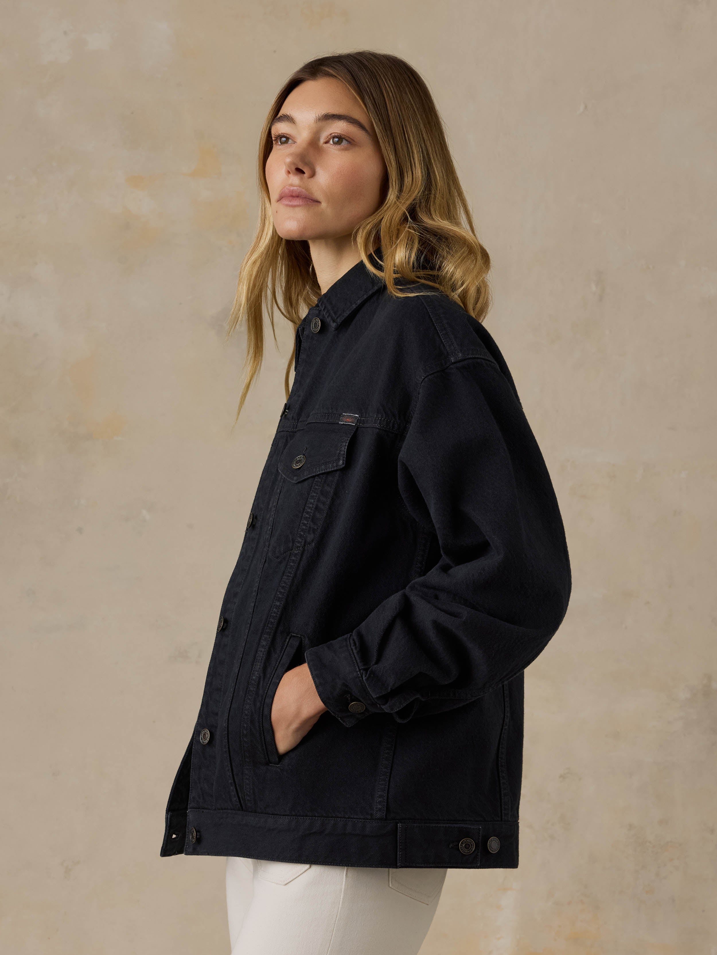 Woman wearing a black denim jacket and white jeans against a beige background