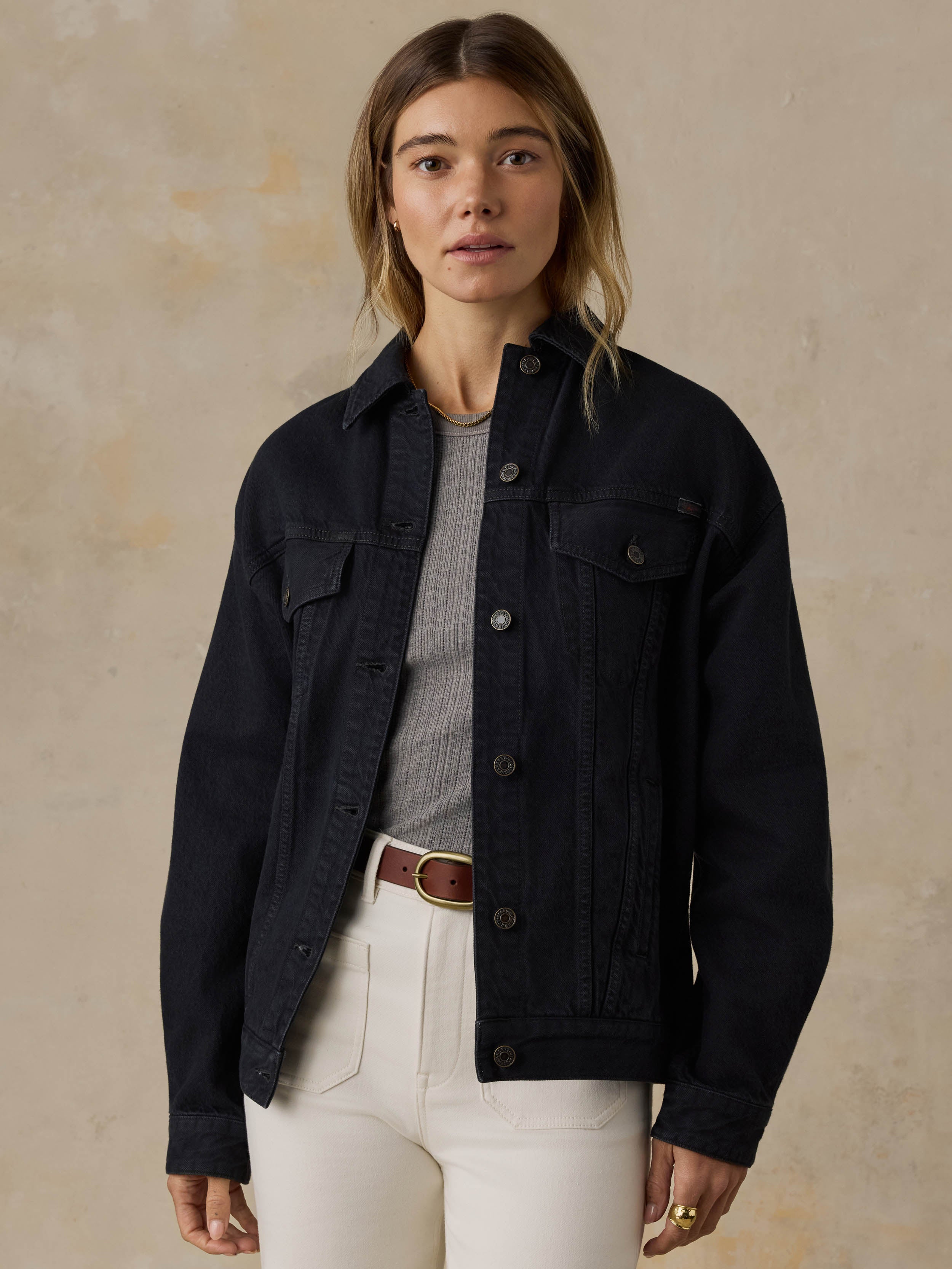 Woman wearing a black denim jacket and white jeans against a beige background