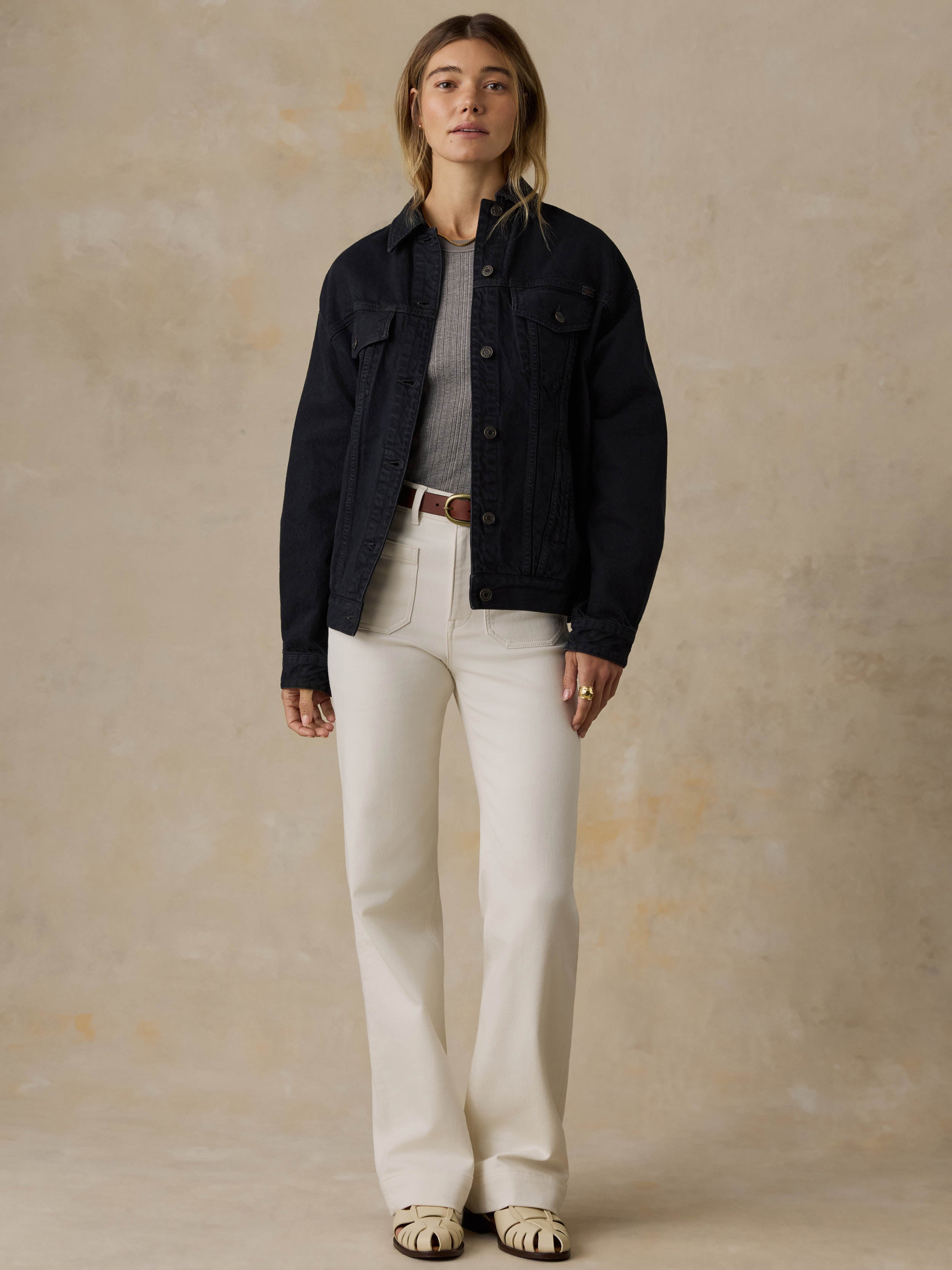 Woman wearing a black denim jacket and white jeans against a beige background