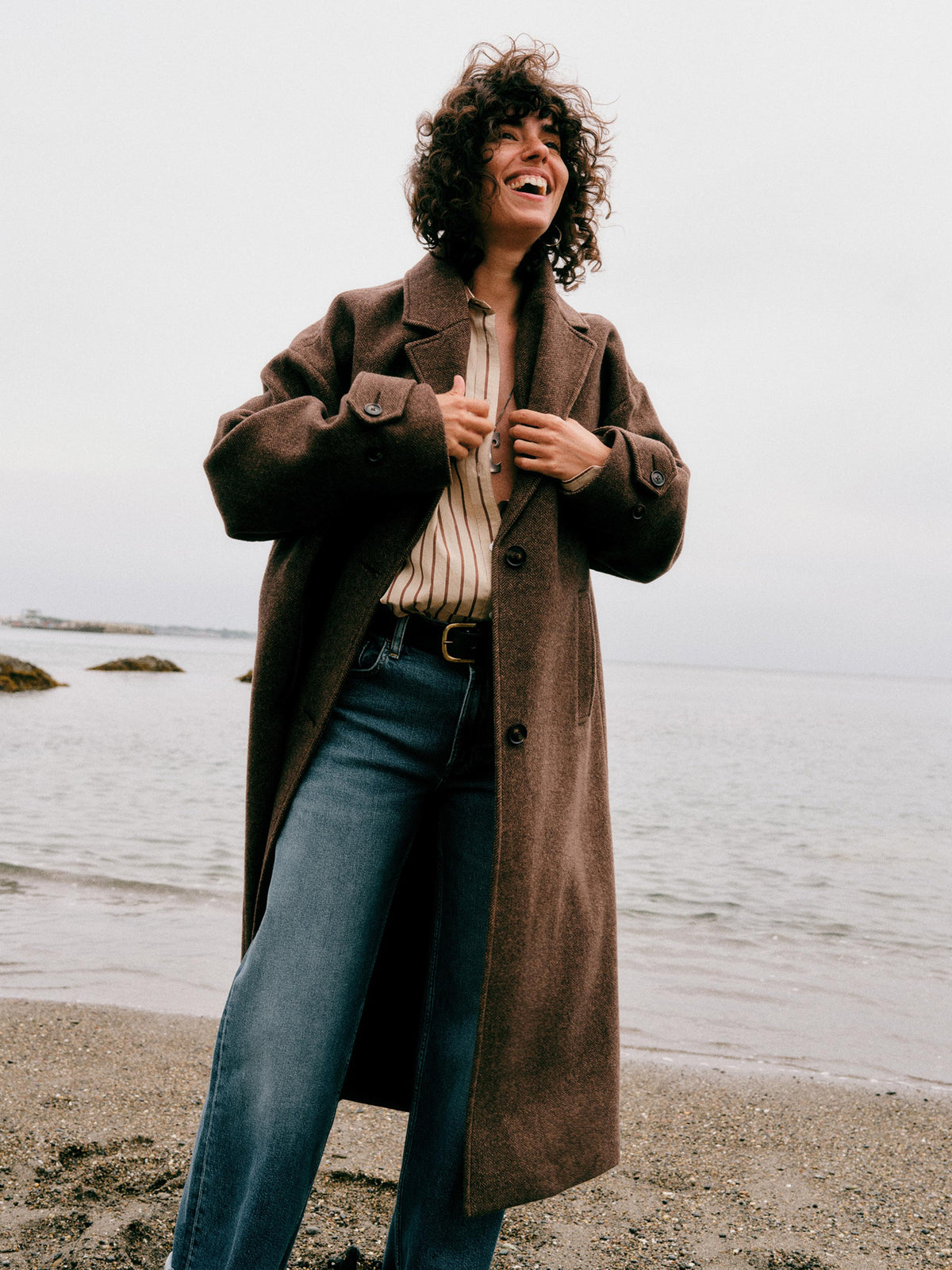A woman wearing a brown coat over a striped shirt and blue jeans, image 1 of 9