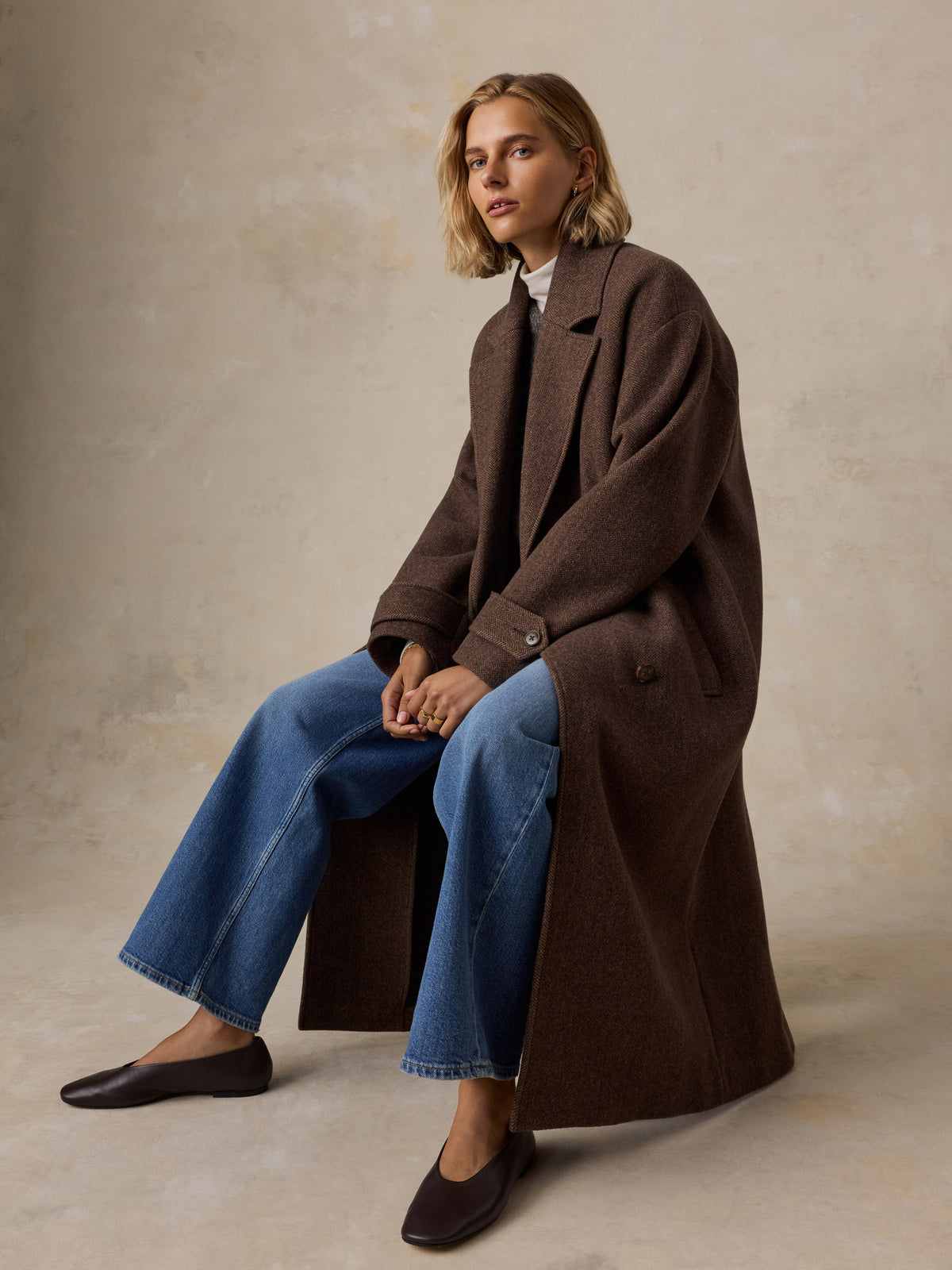 A woman sits wearing a brown long coat over a sweater and blue jeans, image 8 of 9
