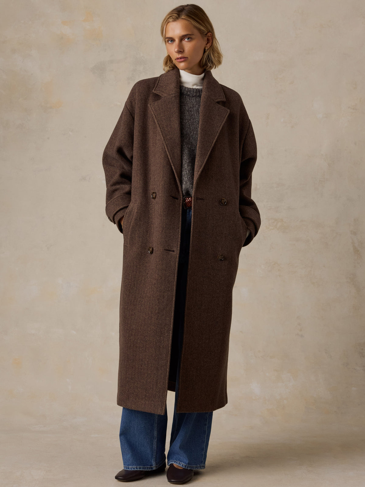 A woman wearing a brown long coat over a sweater and blue jeans, image 2 of 9