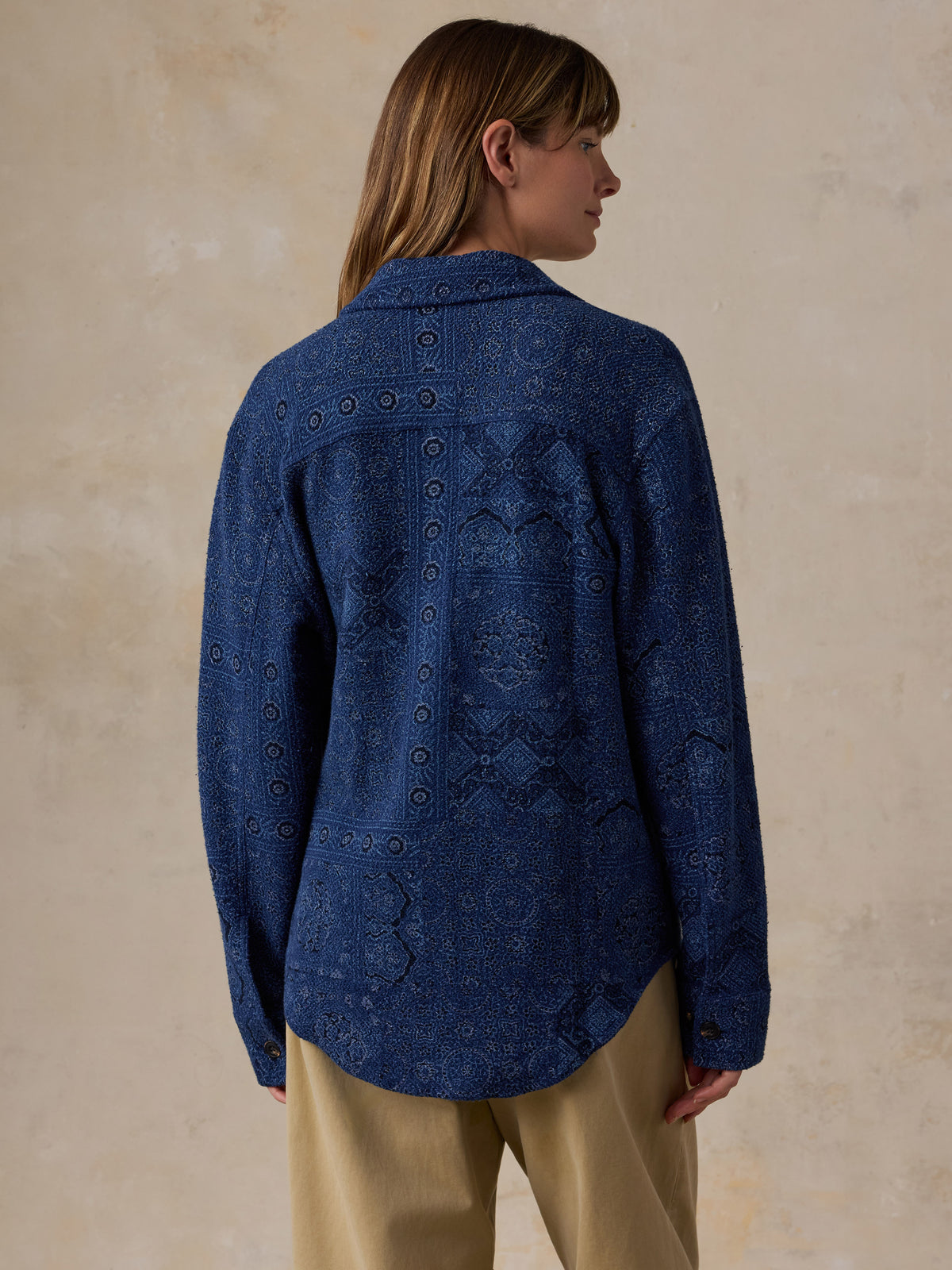 A woman shown from the back wearing a dark blue patterned button-up shirt and beige pants, standing against a textured beige wall., image 4 of 7