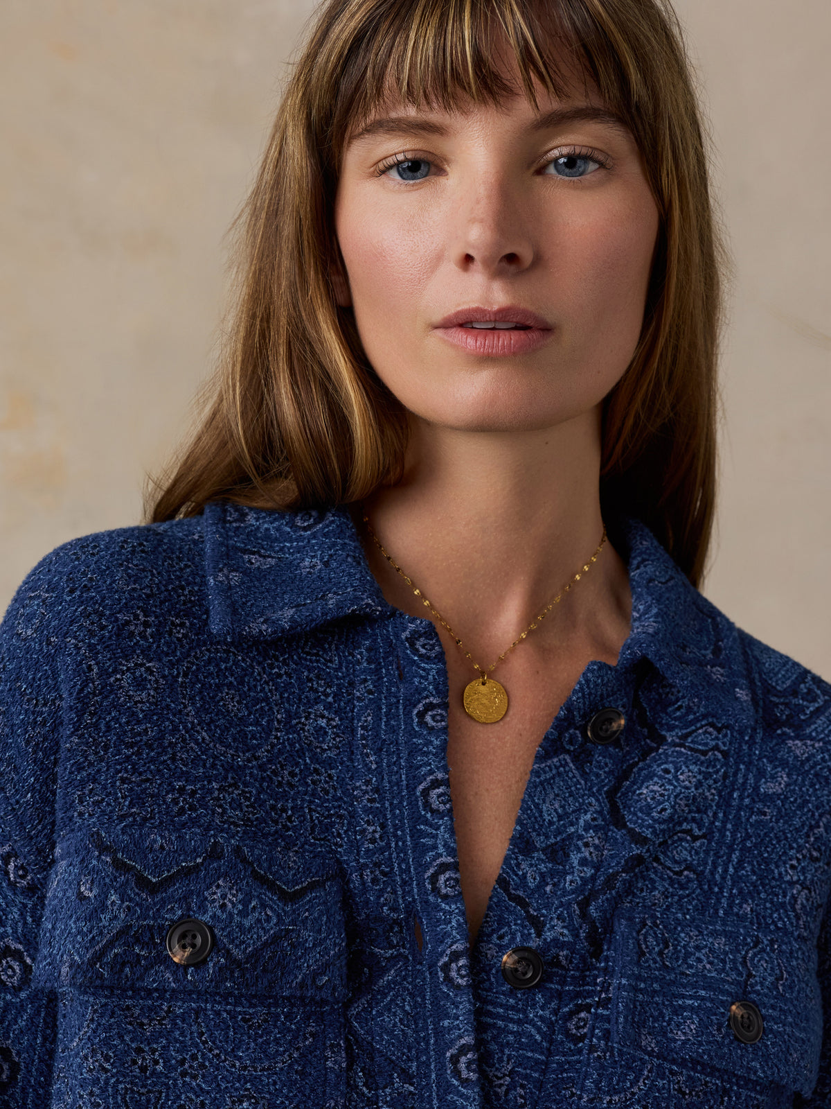 A close-up portrait of a woman wearing a textured blue button-up shirt with a subtle paisley pattern and a gold coin pendant necklace, set against a neutral beige background., image 3 of 7