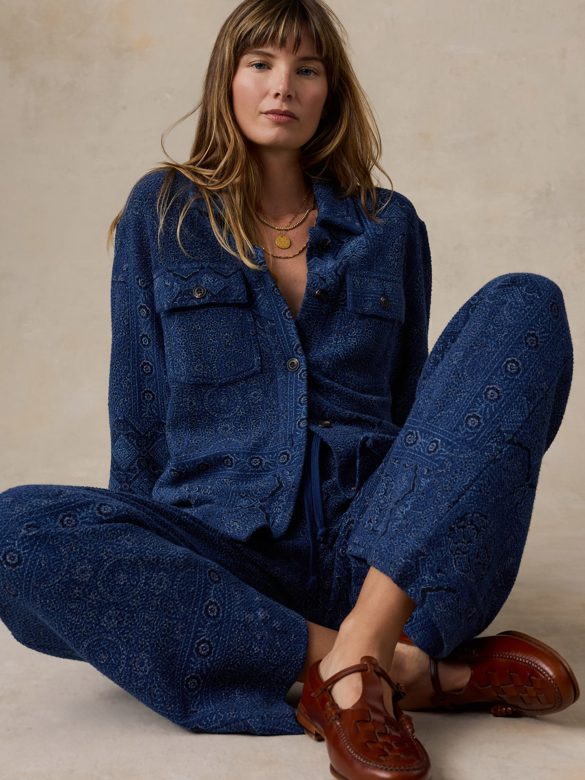 A woman sits cross-legged wearing a matching blue patterned button-front shirt and wide-leg pants set, styled with brown leather Mary Jane shoes against a warm beige textured studio backdrop., image 5 of 7