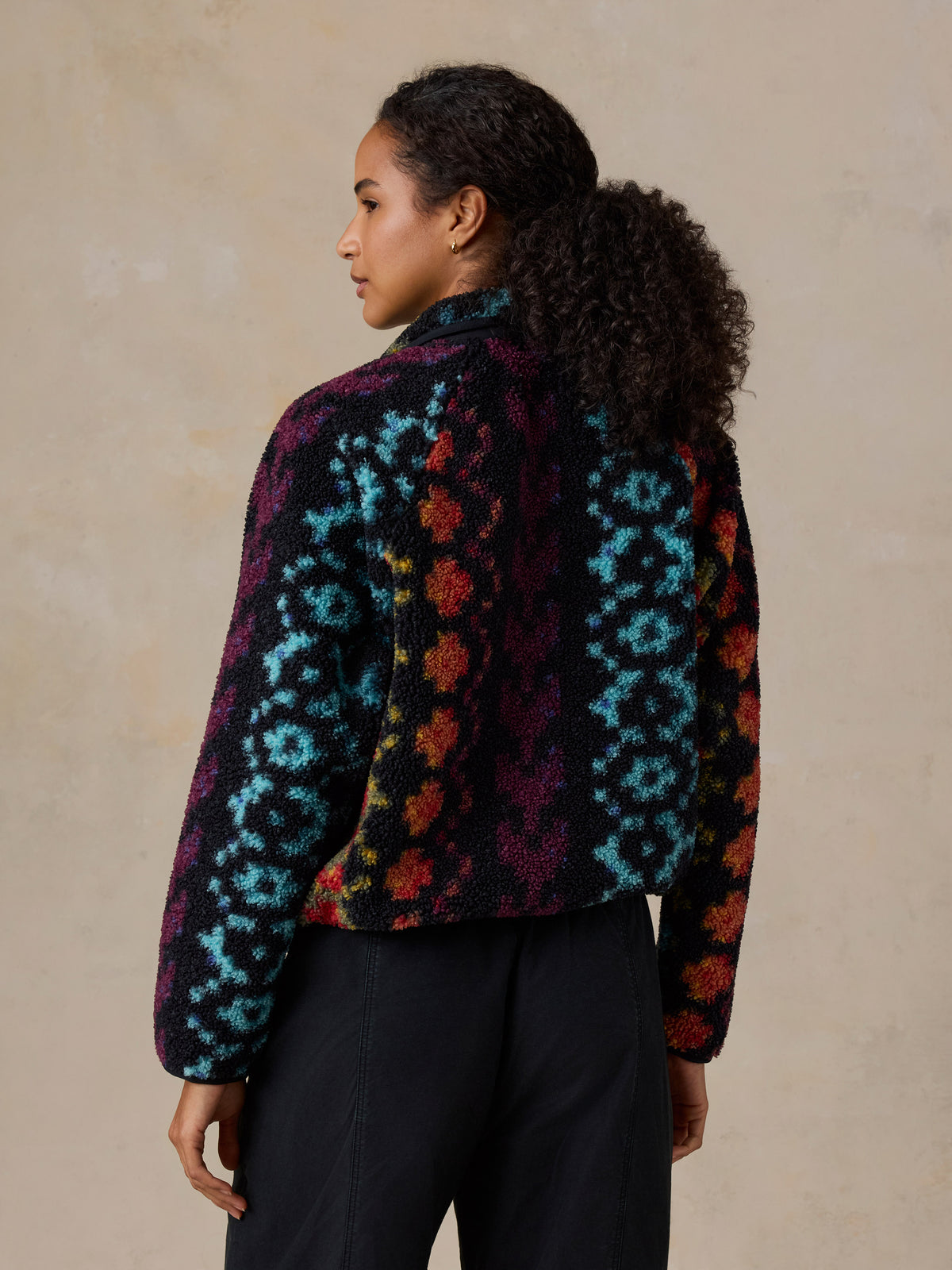 A woman viewed from behind wearing a colorful, patterned fleece pullover and black drawstring pants, image 5 of 7