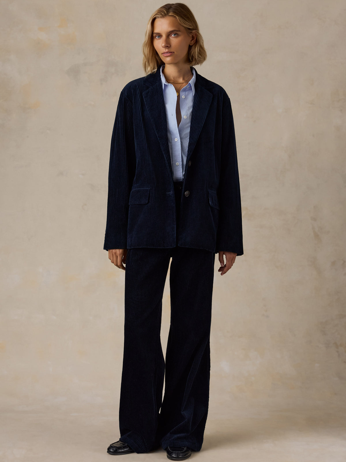 A woman wearing a dark navy blue corduroy suit with a light blue shirt and black shoes, image 7 of 7