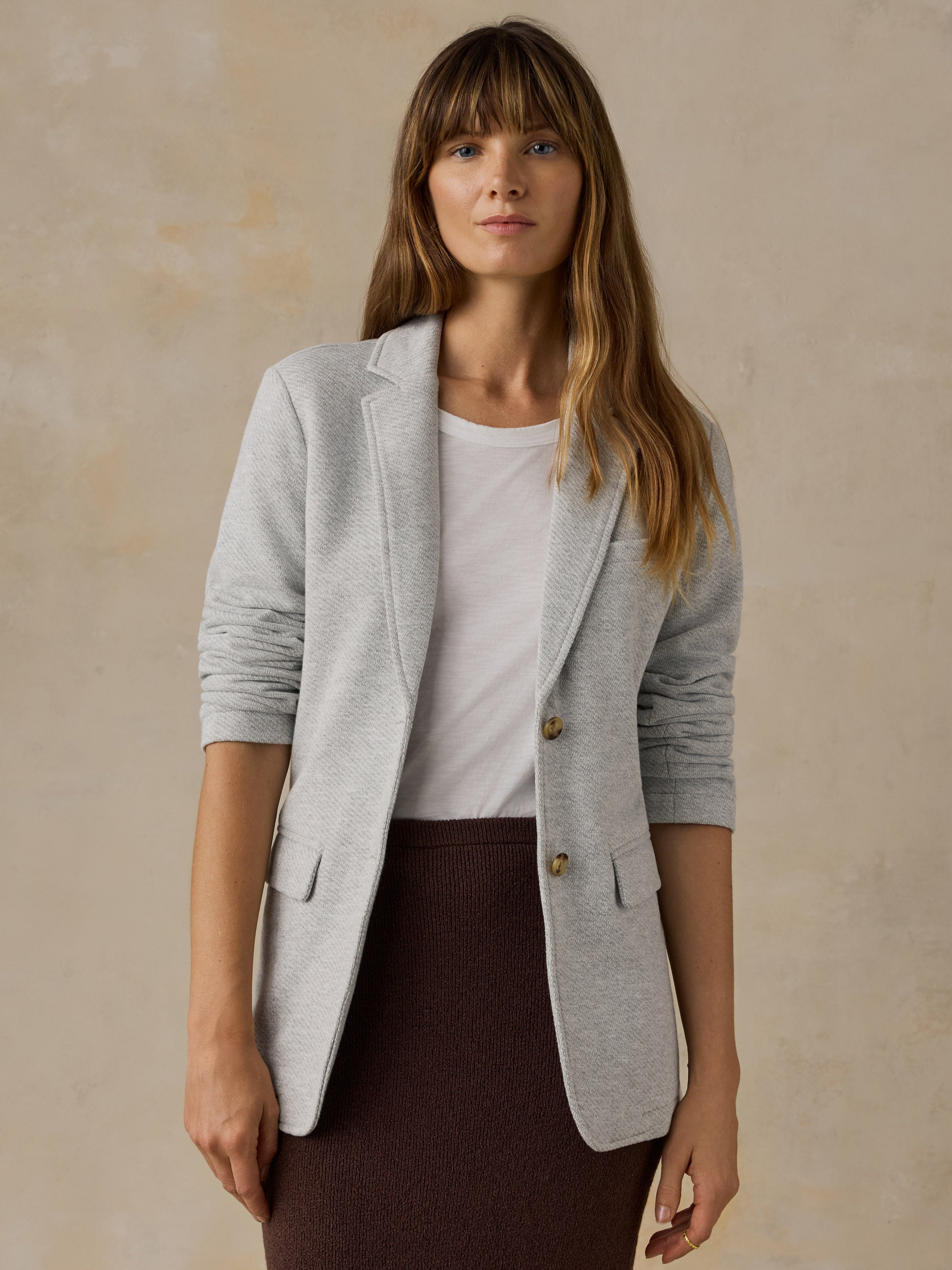 Woman wearing a grey blazer over a white t-shirt and brown skirt against a beige background